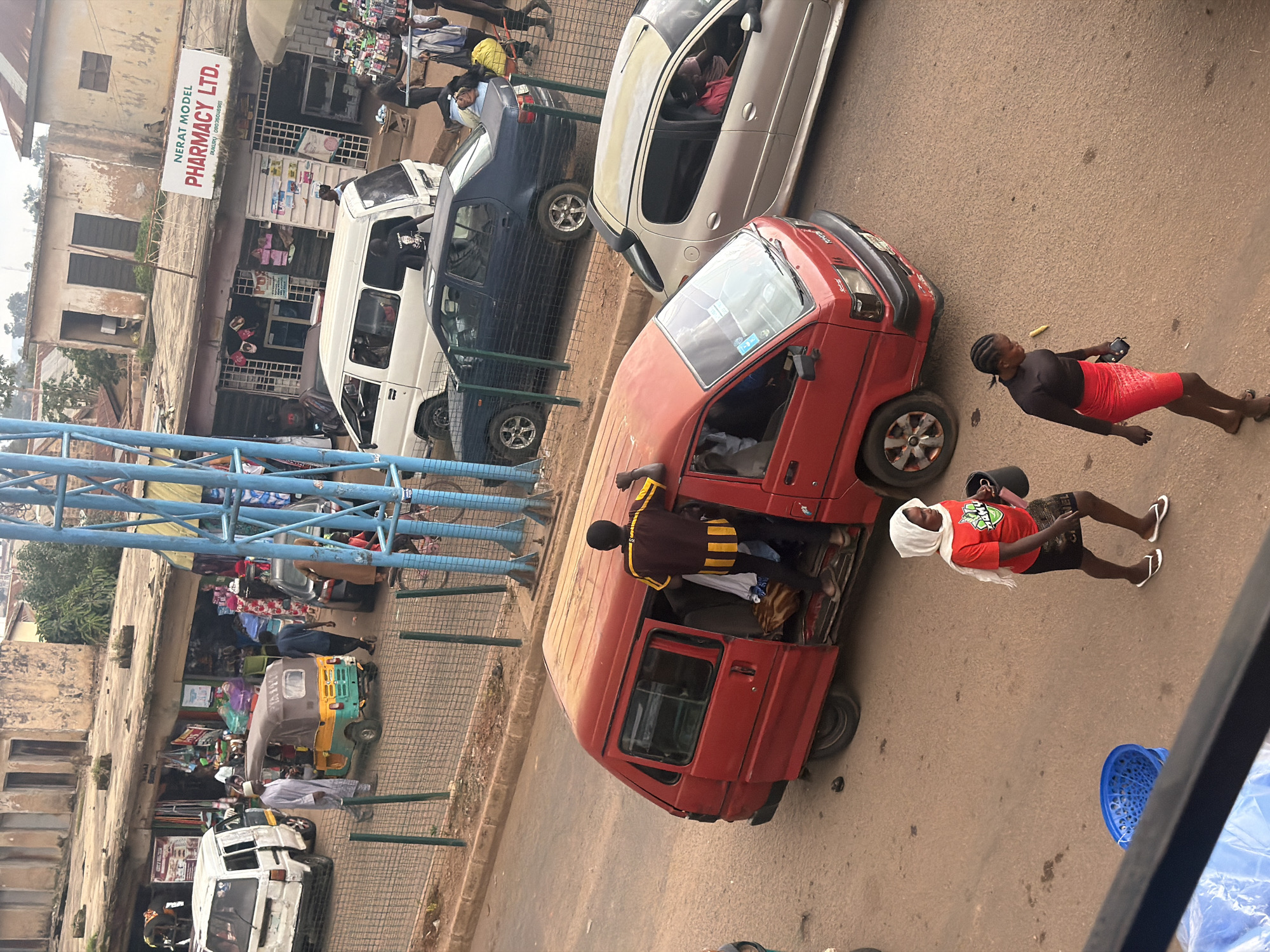 The image depicts a street scene in Bukuru, Nigeria. In the foreground, a red minivan with its doors open is parked, and a person wearing a yellow and brown striped shirt is standing on the threshold. Two women are walking on the street, one in a red t-shirt and white headscarf carrying a black bucket, and the other in a black top and red skirt. A blue plastic basket is on the ground to the left. Behind the minivan, a silver car is parked. Further back, a dark sedan is on the road, and a white minivan is parked. A tuk-tuk is also visible. The background shows buildings with shops, including one with a sign that reads "NERAT MODEL PHARMACY LTD." The street appears to be bustling with activity.