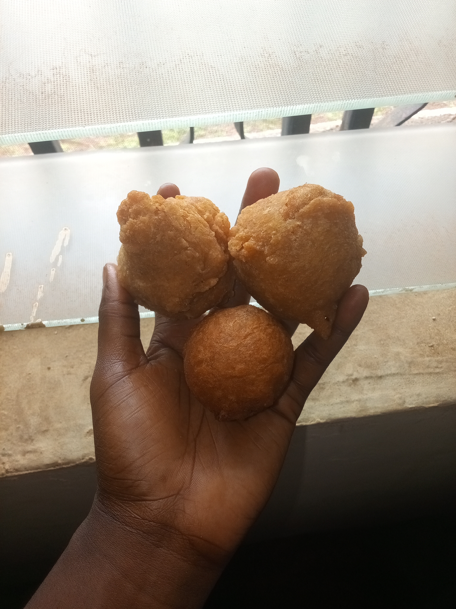 A person's dark-skinned hand is holding three pieces of fried dough against a blurred background of what appears to be a window with a sheer curtain. The fried dough pieces are golden brown and vary in shape and size. One piece is a round ball, while the other two are irregular, puffy shapes. The hand is positioned in the center of the frame, with the fried dough resting on the palm and fingers. The background is out of focus, but suggests an indoor setting with natural light filtering through the window. The overall lighting is soft, casting subtle shadows on the hand and the fried dough.