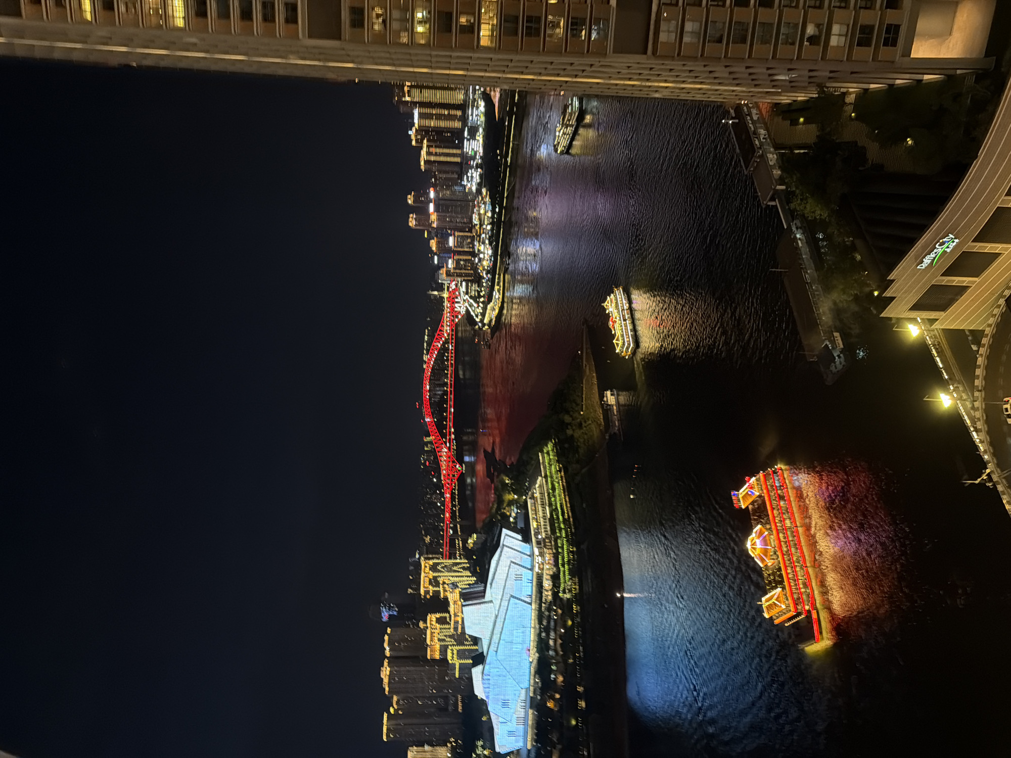 An aerial night view captures a wide river in Yuzhong District, China, with a city skyline illuminated by countless lights. On the left, a large, modern building with blue lighting sits prominently on the riverbank. Across the river, a red arched bridge spans the water, connecting to a cityscape of towering buildings with yellow and white lights. Several boats are visible on the river: a large, multi-decked boat with ornate red and gold lighting is in the foreground, a long, dark boat is near the middle of the river, and two smaller, illuminated boats are further in the distance. The dark water reflects the city lights. On the far right, the side of a tall, modern building with lit windows dominates the frame. The sky is dark and clear.