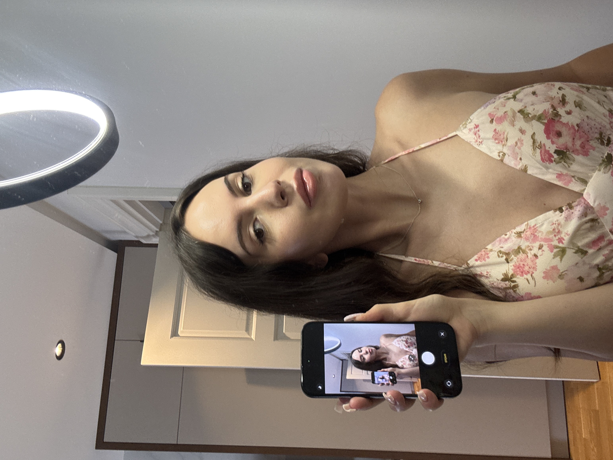 A woman stands indoors, holding a smartphone that captures her reflection in a mirror. The woman has long, dark hair and is wearing a floral-print dress. The phone's screen shows a smaller image of the woman, also holding a phone, creating a fractal effect. Behind the woman, a ring light is visible, suggesting the photo was taken for social media. The background includes a door and what appears to be cabinetry, indicating a domestic setting like a bedroom or bathroom. The lighting is bright and even, consistent with artificial lighting from the ring light and possibly overhead lighting. The overall image is a selfie taken within a reflective surface.
