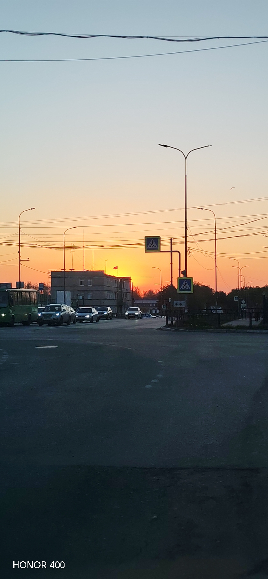 The image depicts an intersection in Chaikovsky, Russia, during a sunset. Several cars and a bus are present on the roadway. A multi-story building with antennae and a flag on top is visible against the setting sun. Streetlights and pedestrian crossing signs are positioned around the intersection. Overhead power lines are strung across the sky. The bottom of the image has the "HONOR 400" logo.