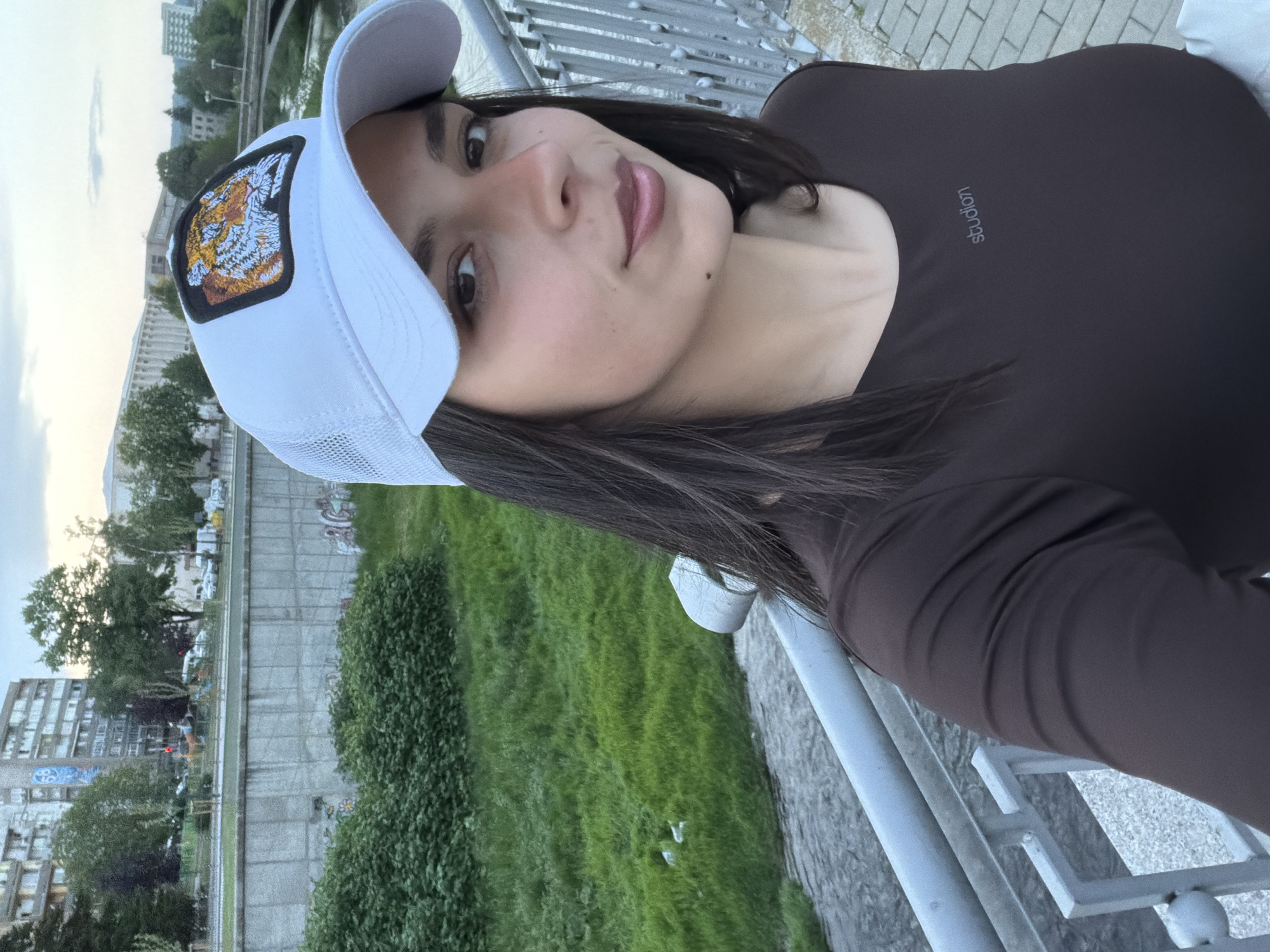 A woman stands on a bridge in Gabrovo, Bulgaria. She is wearing a brown long-sleeved shirt and a white baseball cap with a tiger patch. She is looking at the camera with a slight smile. The background includes a concrete riverbank with green vegetation and buildings in the distance. The metal railing of the bridge is visible in the foreground.