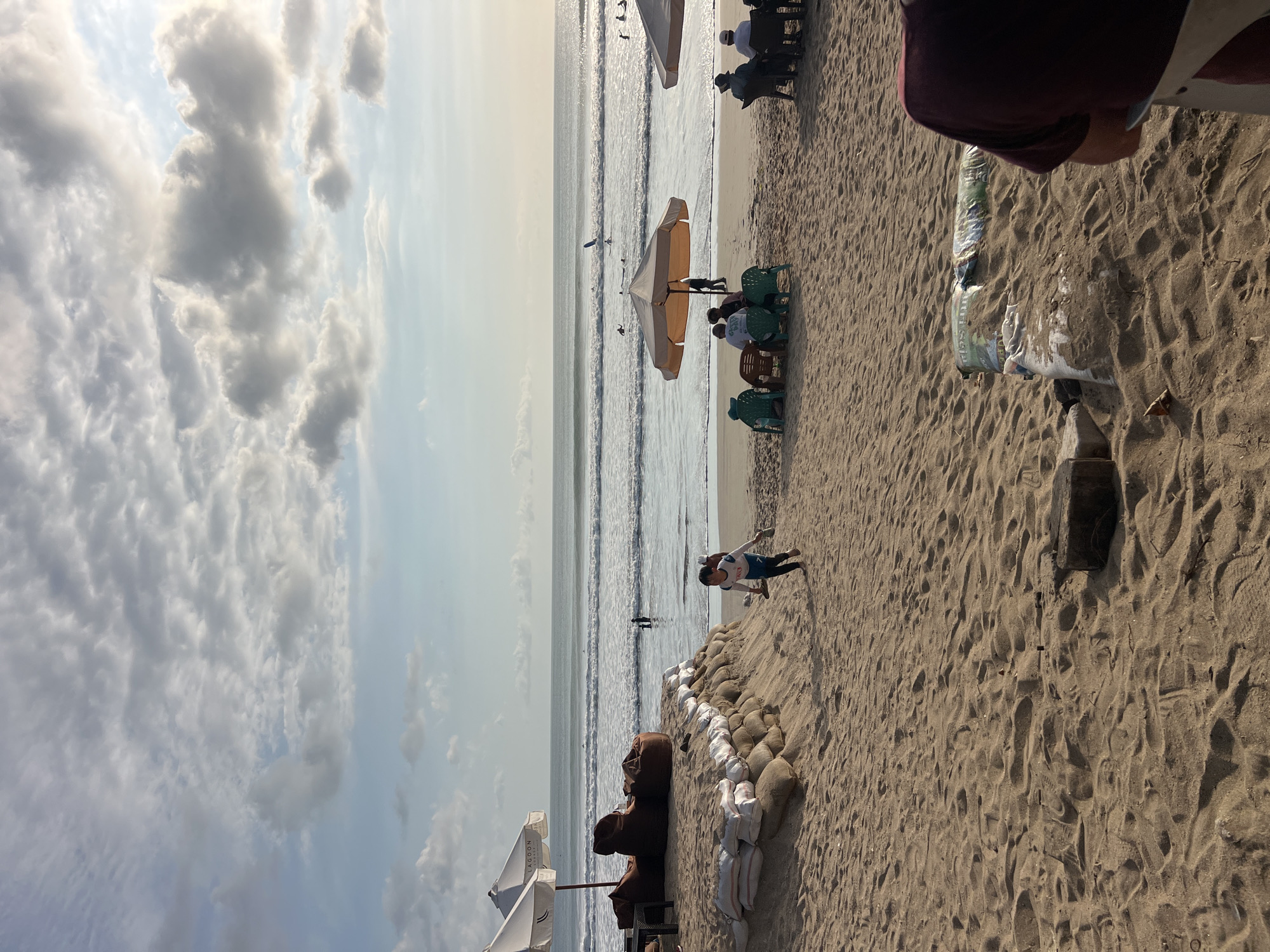 A wide shot of Kuta Beach in Bali, Indonesia, at dusk shows a wide, sandy beach with gentle waves. Several people are in the water, some appearing to be surfers. On the shore, there are lounge chairs and umbrellas for beachgoers. A group of people are seated under a large umbrella, enjoying the view. In the foreground, a child runs along the sand, holding flip-flops. Sandbags are piled up, possibly for erosion control. The sky is filled with dramatic clouds, tinged with the warm light of the setting sun.