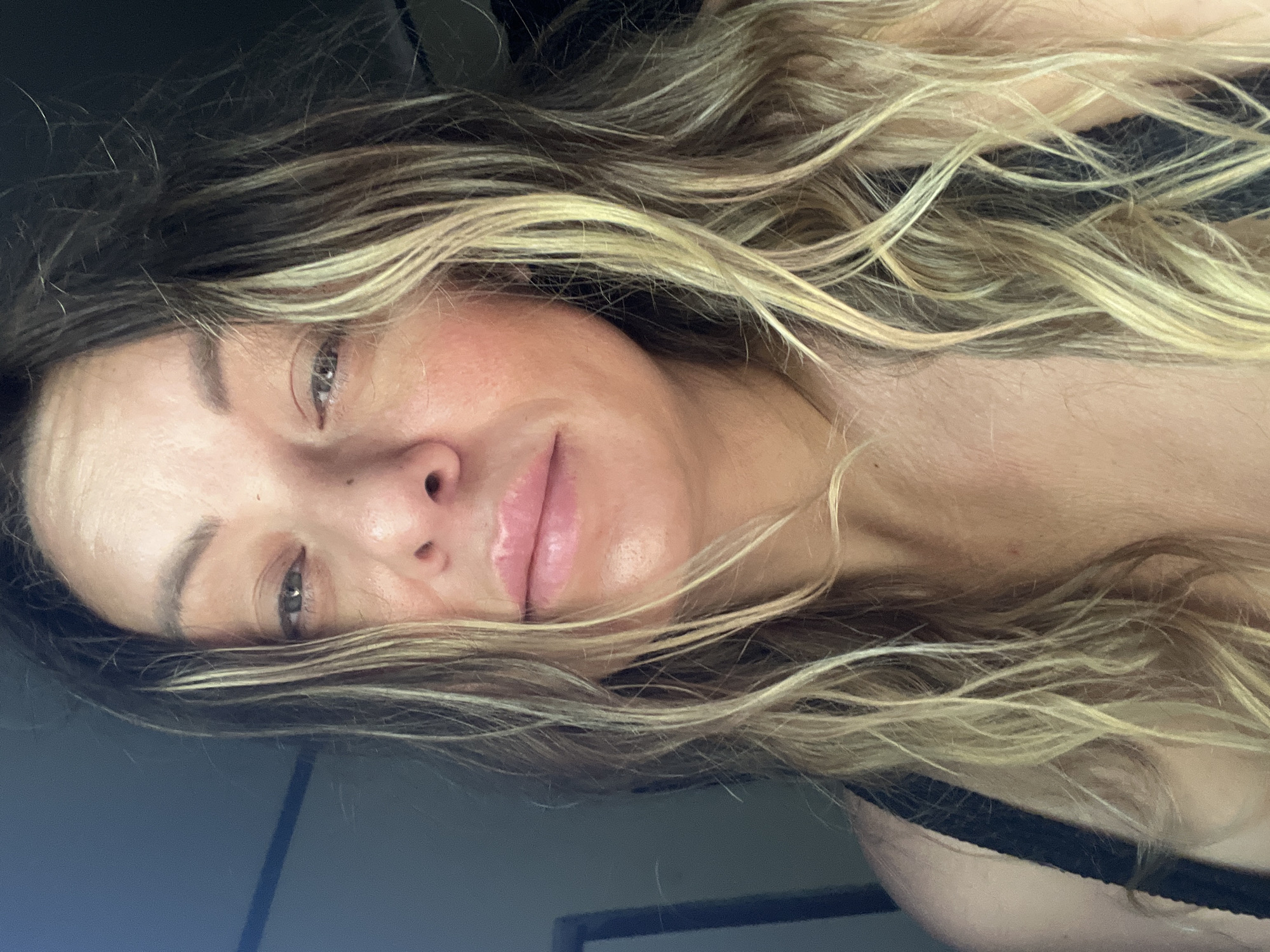 The image is a close-up selfie of a woman with long, wavy blonde and brown hair. She is wearing a black bra. Her face is tilted slightly to her left, and she is looking directly at the camera with a neutral expression. Her skin appears to be smooth with some subtle blush on her cheeks. The background is dark and out of focus, with a hint of a blue-grey wall on the left side of the frame. There are no identifiable landmarks or details that would suggest a specific location within a city. The lighting appears to be soft and natural, possibly from a window.