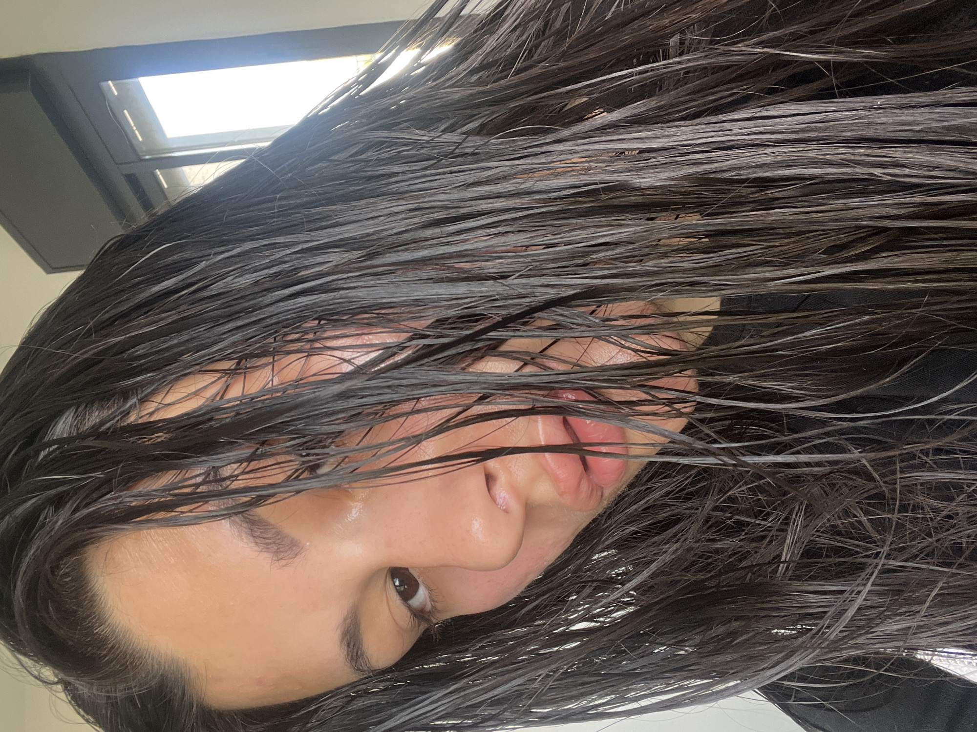 A close-up, rotated image shows a person with wet, dark brown hair partially obscuring their face. The person has light skin, dark eyebrows, and visible facial features including an eye looking left, a nose, and slightly parted lips. A rectangular light source or window is visible in the upper left corner. The setting appears to be indoors. The location is not apparent from the image. No actions or interactions are visible. The exact location of Kiryat Bialik, Israel, is indeterminable from this image.