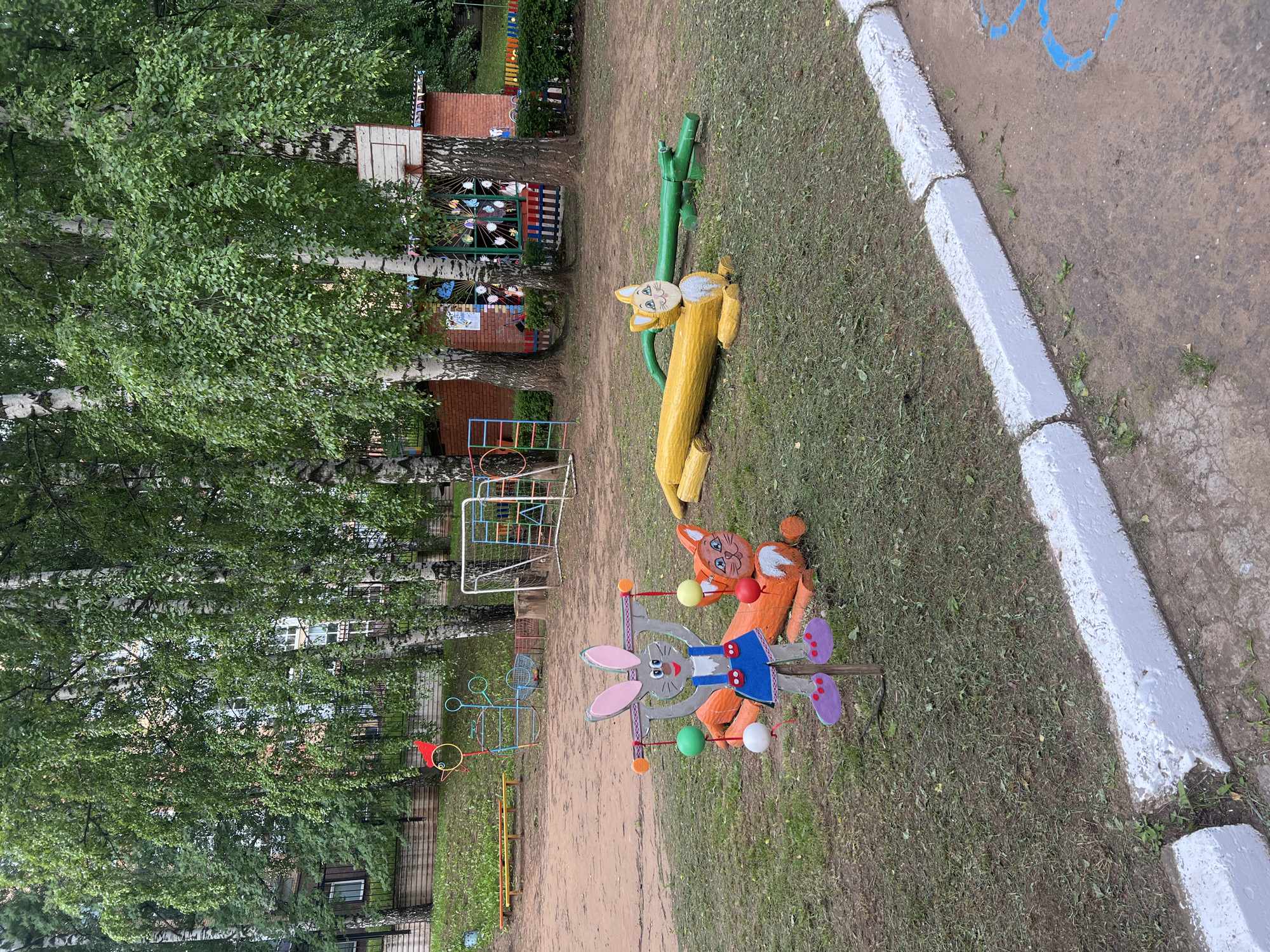 In the city of Sovetsko-Nikolskoye, Russia, an outdoor playground scene is depicted. The foreground features a whimsical wooden sculpture of a rabbit holding colorful balls, standing in front of a carved orange fox sculpture. Further back, a yellow log animal and a green crocodile-like sculpture are visible on the grass. Behind these, a dirt area is marked by a low white concrete curb. The background is dominated by birch trees with lush green foliage, casting dappled shade. A brick building with windows and a basketball hoop is partially visible amongst the trees, along with playground equipment like a soccer goal and a climbing frame. The overall impression is one of a well-used, if slightly worn, children's play area.