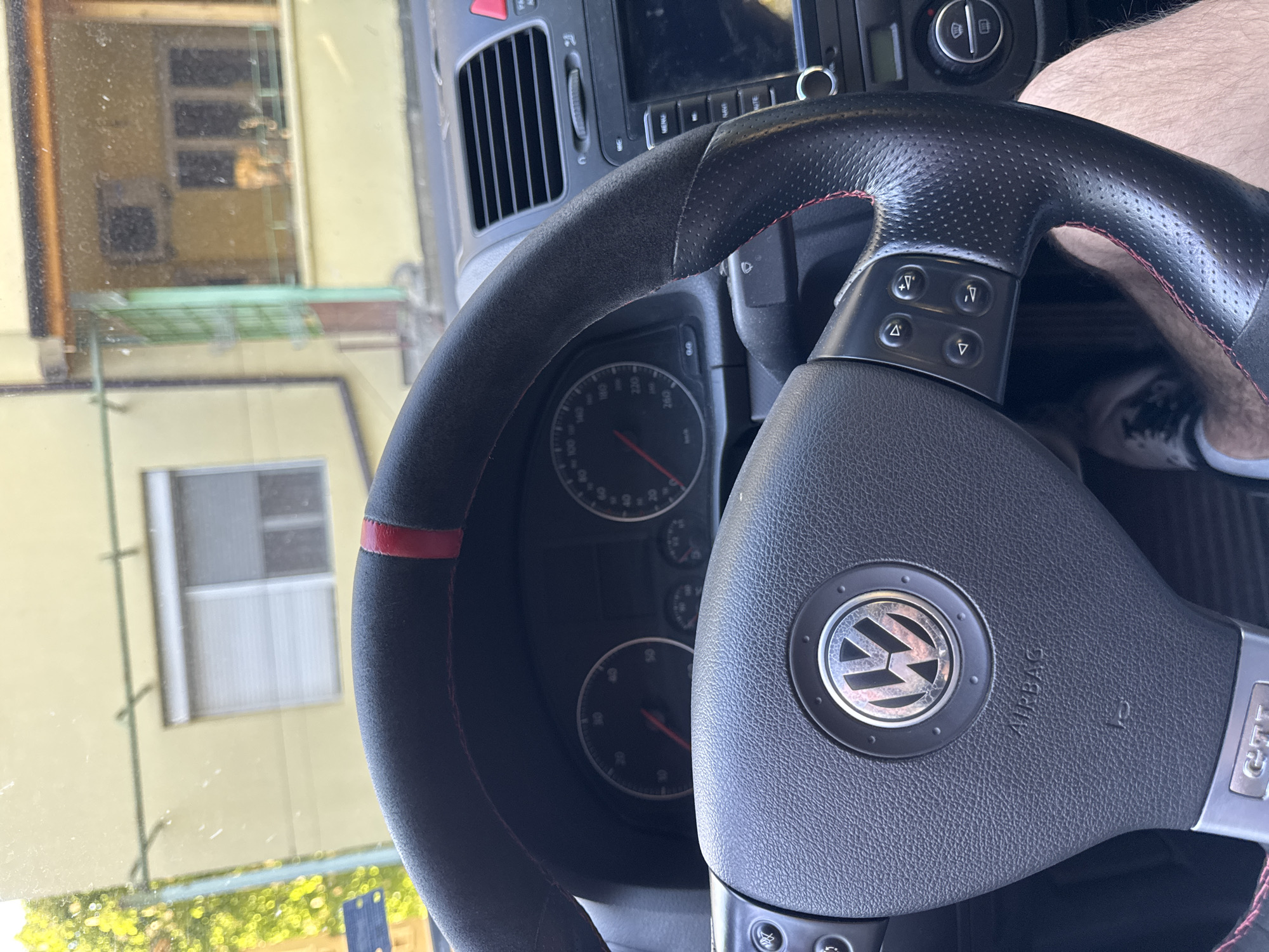 A close-up shot from inside a car focuses on a black steering wheel with a Volkswagen logo and a "GTI" badge. The wheel has a suede-like material on the outer rim with a red stripe at the top. Buttons are visible on the steering wheel, likely for controlling various car functions. The dashboard with speedometer and other gauges is also in view. Through the car's windshield, a building with yellow-painted walls and windows can be seen. The location appears to be a residential area, likely in Pleven, Bulgaria, based on the urban architectural style. A person wearing a shoe is visible behind the steering wheel.