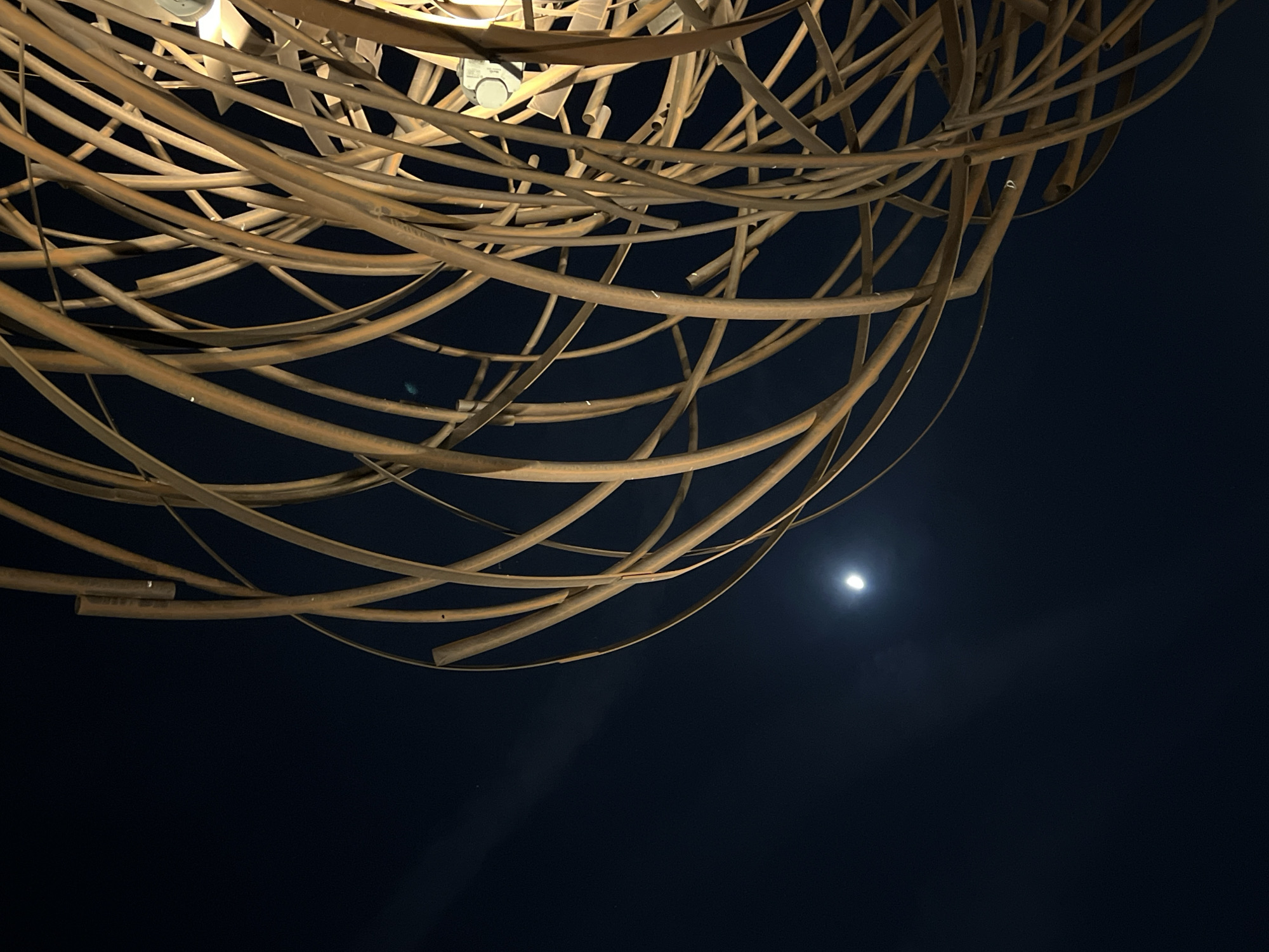 The image is a low-angle shot taken at night. In the upper portion of the frame, there is an intricate, spherical structure made of curved, metallic, rod-like elements. A light source is visible at the top center, likely illuminating the structure. The bottom portion of the image is dominated by a dark night sky, with a small, diffuse, bright spot that could be the moon or a distant light source. The scene appears to be urban, given the artificial light and the constructed object.
Based on the presence of the sphere made of rods, it is possibly located nearby the Palacio de Cristal, in the Retiro Park, in Madrid, Spain, where this structure can be seen. However, this can not be inferred for certain.