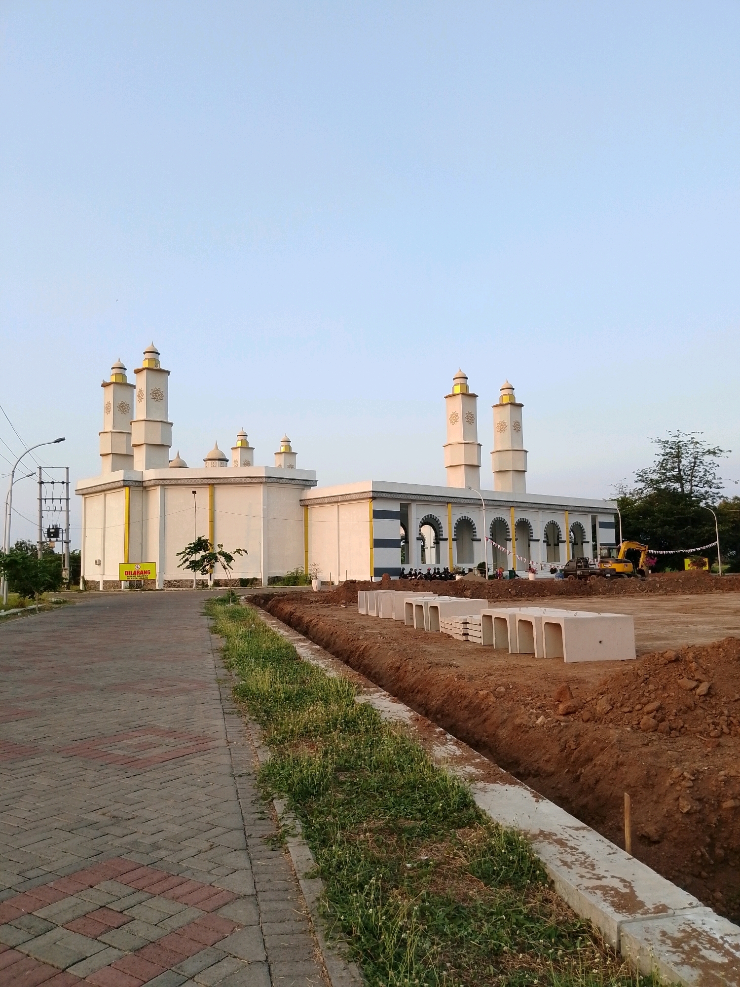 The image shows a modern mosque with white walls and golden domes, located in Pasuruan, Indonesia. The mosque has tall minarets and a series of arched entrances. In the foreground, a paved walkway leads towards the mosque, bordered by a grassy area and a trench. Construction materials, including concrete structures, are visible in the foreground, suggesting ongoing development or renovation. A yellow construction vehicle and a pickup truck are parked near the mosque. The sky is clear and blue, indicating daylight. The scene appears to be an outdoor public space.