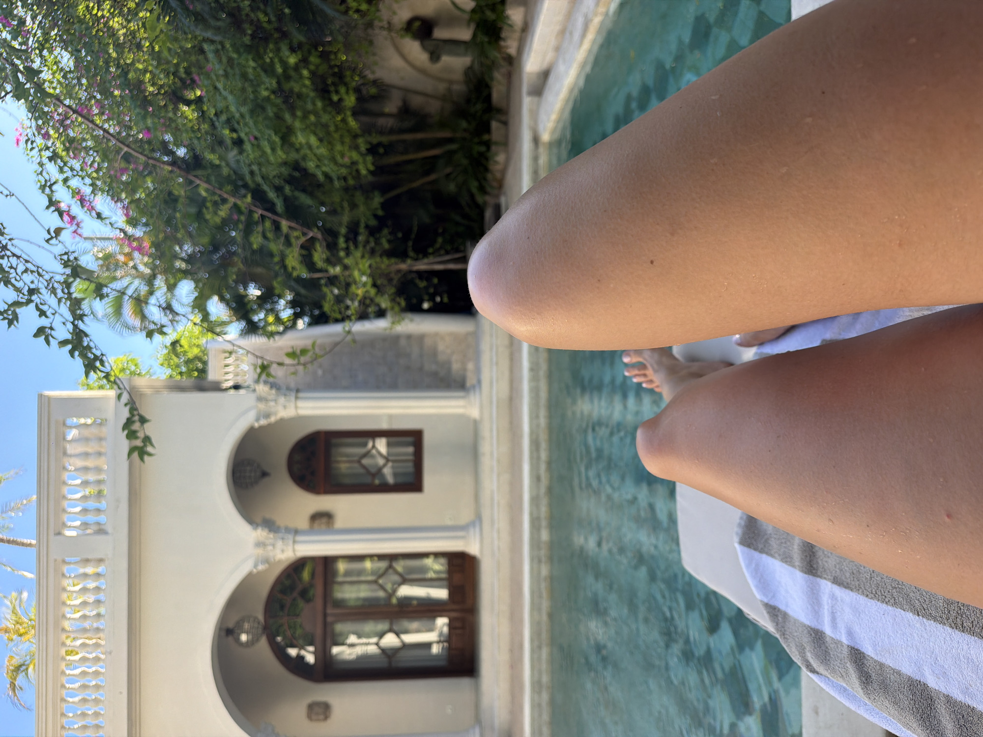 The image captures a first-person perspective of someone relaxing by a swimming pool in a tropical resort setting. In the foreground, the person's tanned legs and feet are visible, resting on a lounge chair covered with a white, grey, and black striped towel. Small water droplets on the skin suggest recent time spent in the water.
The turquoise swimming pool extends from the mid-ground to the right side of the frame. Beyond the pool, a grand white building with Mediterranean-inspired architecture dominates the background. It features multiple arched doorways and windows with dark wooden frames and intricate diamond-shaped glass panes. Decorative columns support the structure, and an ornate white balustrade runs along a balcony or upper level.
Lush green vegetation, including palm trees and flowering vines with bright pink blossoms, frames the upper and left portions of the image against a clear blue sky. The scene is brightly illuminated by natural sunlight, indicating it is daytime. The overall ambiance conveys relaxation and luxury in a warm, tropical environment, consistent with North Kuta, Indonesia. No visible text is present in the image.