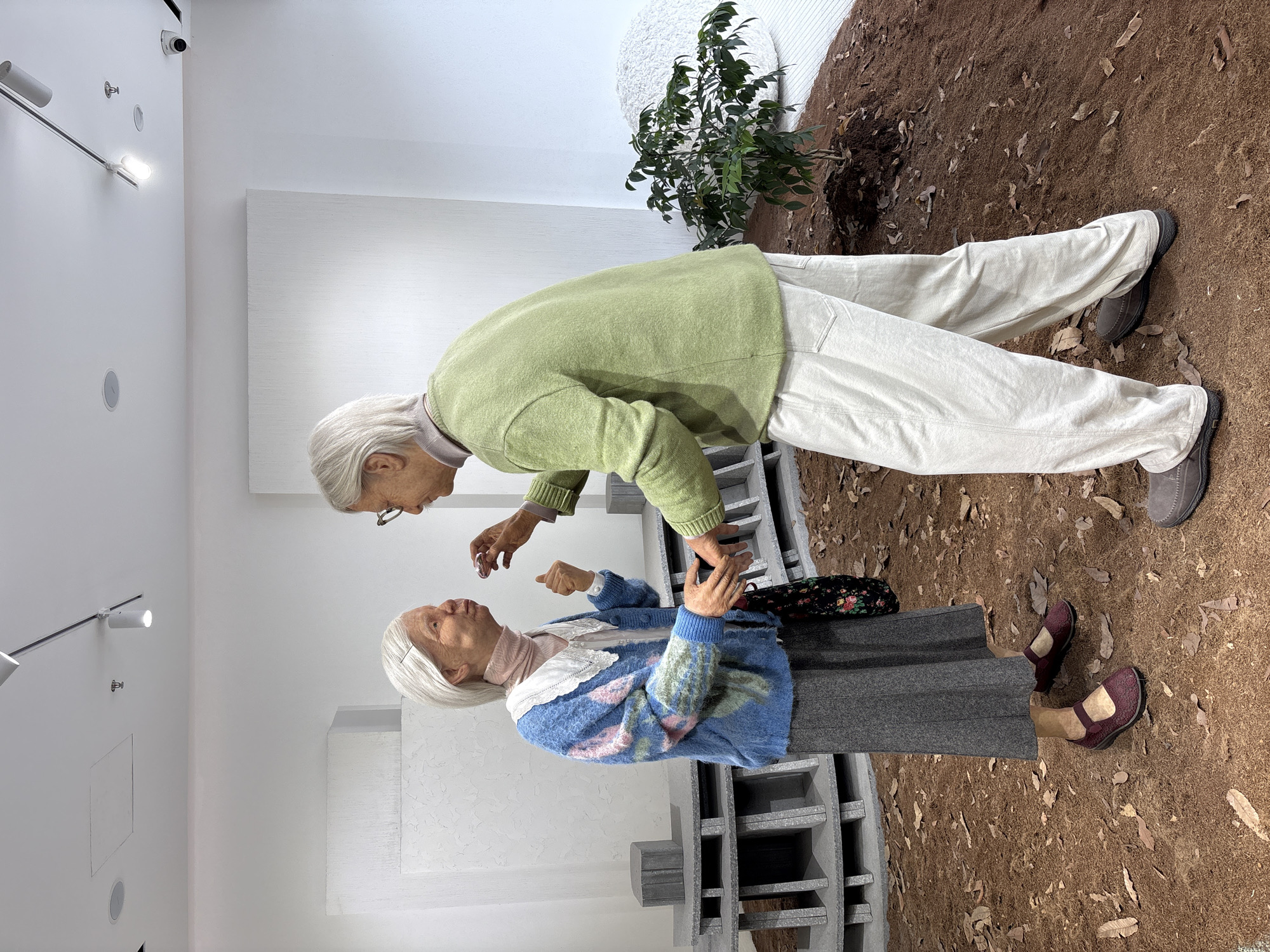 This is a photograph of two lifelike wax figures of elderly people in an indoor exhibition space. The figures are positioned facing each other in a way that suggests a conversation or interaction. The figure on the left is a woman wearing a blue knitted cardigan with floral patterns, a light pink turtleneck, and a grey skirt. She has white hair pulled back and is holding a small dark object, possibly a piece of candy or medication, up to her mouth with her left hand. Her right hand is extended towards the figure on the right. The figure on the right is a man with white hair and glasses, wearing a light green crewneck sweater and loose, light-colored pants. His left hand is extended towards the woman, and his right hand is reaching out to her. The figures are standing on a ground surface covered with brown mulch and fallen leaves, designed to resemble an outdoor environment. Behind the figures, there are elements of exhibition decor, including minimalist grey structures that resemble tiered shelving or seating, and a white textured wall. A small green plant is visible to the right of the man. The lighting appears to be from artificial track lighting on the ceiling. The overall presentation suggests a contemporary art installation or museum exhibit. The location is the Shanghai Pudong Museum of Art.