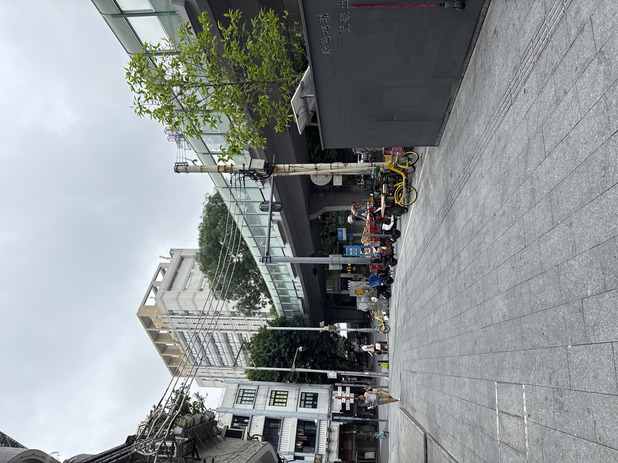 The image depicts a street scene in Yuexiu District, China, characterized by an elevated pedestrian walkway or overpass spanning across the street. The walkway is constructed with a grey base and features a translucent green enclosure with windows. Below the walkway, a series of utility poles and wires are visible.
On the left side of the street, a multi-story building with a light-colored facade and multiple windows stands prominently. Adjacent to it, another building with a darker facade and visible laundry hanging from windows is present. The street itself is paved with grey granite tiles.
In the foreground and midground, several bicycles, electric scooters, and a yellow tricycle are parked along the sidewalk. Some appear to be shared mobility devices. A few individuals are visible walking on the sidewalk. One person in the midground is walking away from the camera, and another is seen near the parked vehicles.
On the right side of the street, a dark grey, opaque structure with some text inscribed on it is visible, topped with a solar panel. The environment appears to be overcast, contributing to a diffused lighting condition. Trees with green foliage are interspersed throughout the scene, particularly near the elevated walkway and in the background. The overall impression is an urban streetscape with a mix of modern infrastructure and everyday city life.