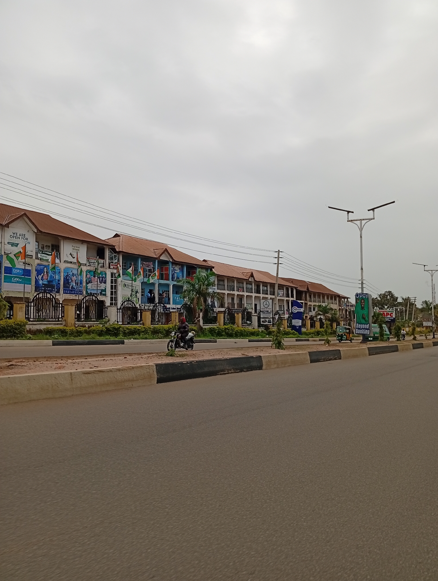 The image shows a street scene in the city of Gura Top, Nigeria. A long, multi-story building with a reddish-brown tiled roof lines the right side of the street. The building appears to be a commercial or residential complex with numerous windows and balconies. Various advertisements and banners are displayed on the facade of the building, including what appears to be an advertisement for "OPay Beyond Banking" and another for "APC". Several streetlights are visible along the street.
In the foreground, a paved road takes up the lower portion of the image. A concrete barrier with black and white markings separates the road from a green strip of vegetation and then the building. A motorcycle with a rider is seen on the road to the left, and a few other vehicles, including a tricycle and a van, are parked further down the street. The sky is overcast and gray, suggesting a cloudy day. The overall impression is a moderately busy street in an urban or semi-urban setting in Nigeria.