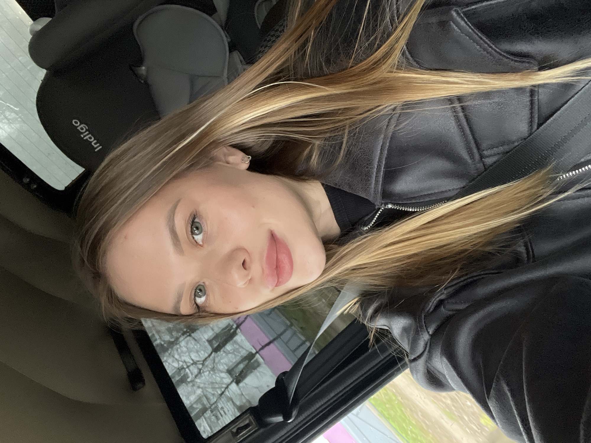 A young woman is captured in a close-up selfie taken inside a car, likely in Chaikovsky, Russia. She has long, straight, blonde and light brown hair that cascades over her shoulders and chest. Her light-colored eyes (grey or green) gaze directly at the camera, and she wears a subtle, pleasant expression with a slight smile on her light pink lips. She is dressed in a black leather-look jacket with a prominent silver zipper, over a dark, presumably black, top. A car seatbelt crosses her shoulder.
In the background, to the left of the woman's head, a dark grey or black child car seat is visible, bearing the white text "Indigo" vertically printed on its fabric. Through the car window to her right, a blurred urban or suburban environment can be seen. This includes indistinct buildings, one of which appears to have pink elements, and bare tree branches suggesting either late autumn or early spring. The lighting indicates it is daytime, possibly overcast, given the soft, diffused light. A small, metallic-colored detail is visible on one of her fingernails. The image itself appears to be originally rotated.