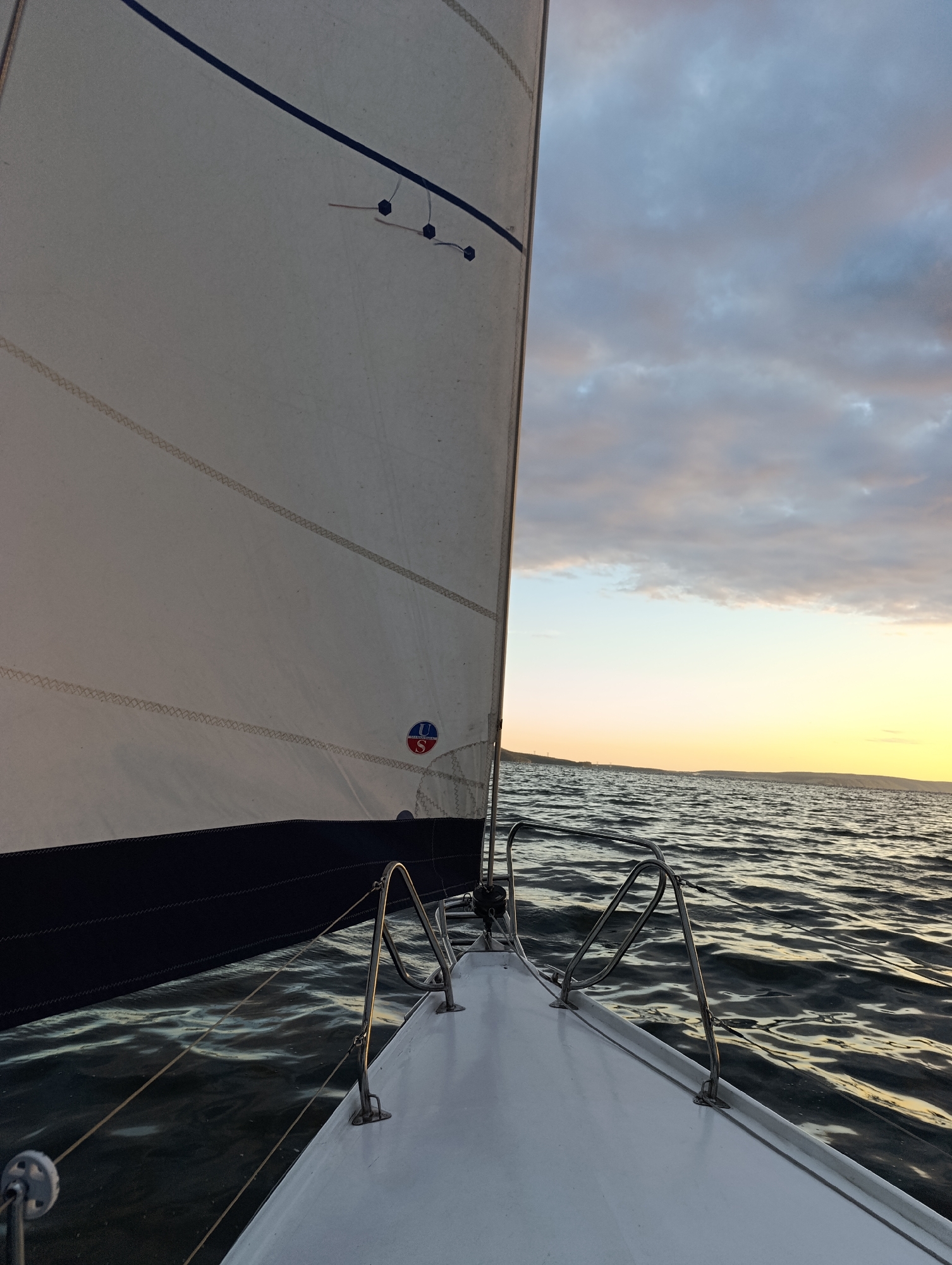 The image shows a sailboat on the water during sunset. The sail is partially visible on the left, with a dark blue stripe along its bottom edge. The bow of the sailboat is in the foreground, with chrome railings. The water is choppy and reflects the warm colors of the sunset sky. The horizon line is visible, with a distant landmass and a soft, yellow-orange glow from the setting sun. The sky is cloudy, with patches of blue and grey, and some areas of pink and orange from the sunset. The location is Tolyatti, Russia, which is on the banks of the Kuybyshev Reservoir, a large artificial lake formed by the damming of the Volga River.