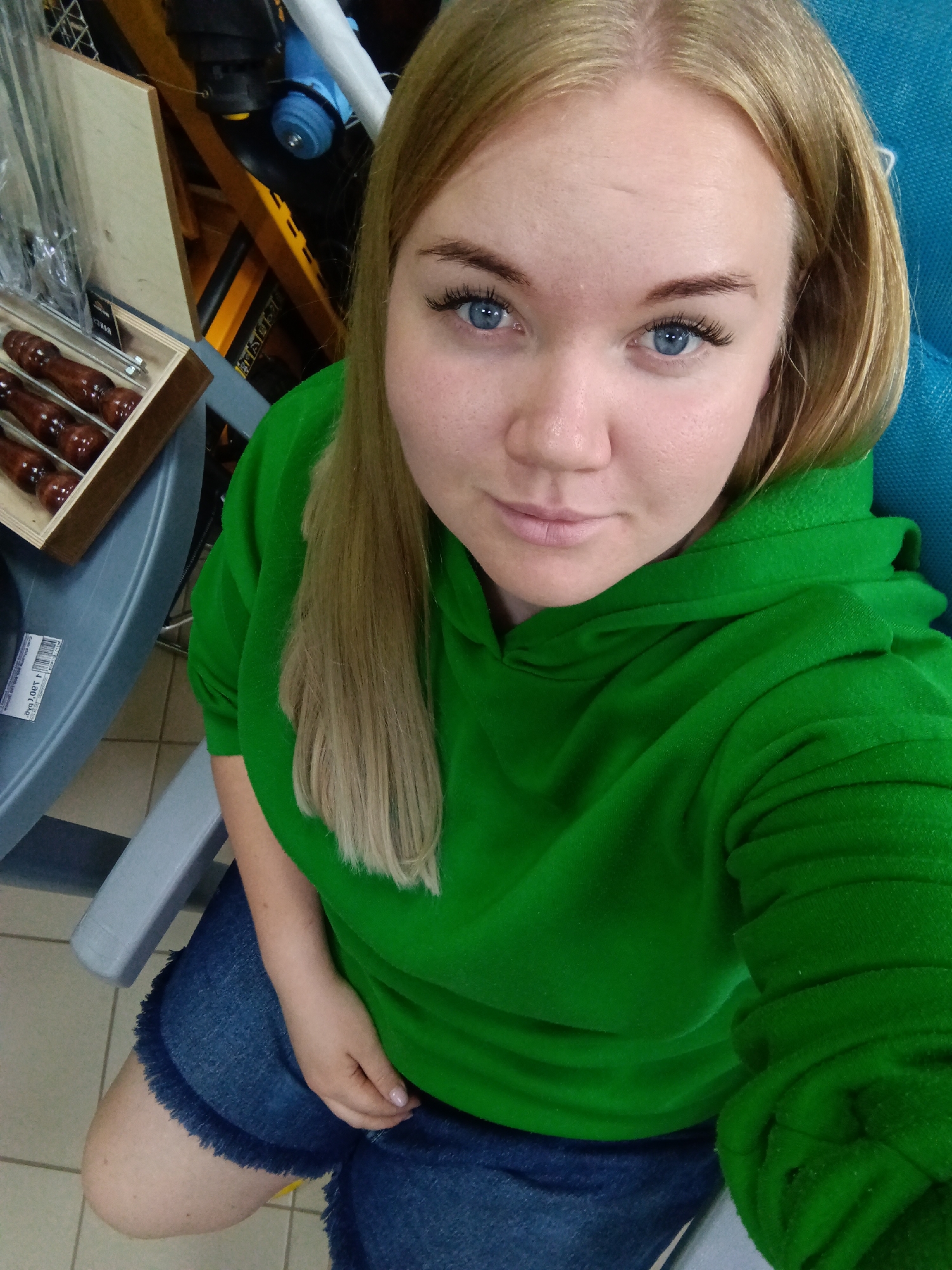 A medium shot captures a young woman with blonde hair and striking blue eyes, gazing directly at the camera. She wears a bright green hooded sweatshirt and blue denim shorts with frayed hems. Her hair is parted slightly off-center and falls over her shoulders. She appears to be seated or kneeling.
To her left, a light grey plastic surface, possibly a table or chair armrest, is visible. On this surface, a wooden box holds several items with dark, polished wooden handles, likely skewers or cutlery, suggesting a retail display. On a white sticker on the grey surface, partially visible and inverted text reads "ow.J.Q£ r.j".
In the background, hints of shelving and various objects in yellow, black, and blue are visible, consistent with an indoor commercial or storage environment, such as a store or market in Фоки, Russia. The floor is tiled with light-colored squares. The woman's expression is neutral and calm as she poses for the photograph.