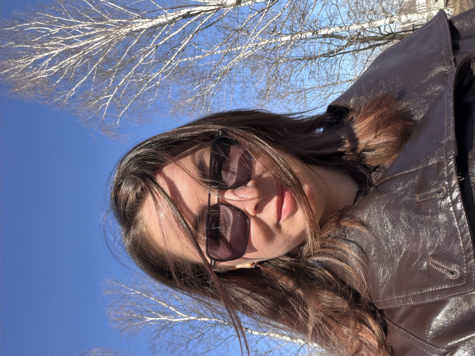 A low-angle portrait shows a woman with long brown hair wearing sunglasses and a brown leather coat. The background features a bright blue sky and bare tree branches. While the location cannot be determined from the image alone, the prompt requests that it be in Izhevsk, Russia. Therefore, the image may be said to depict a woman in Izhevsk, Russia.