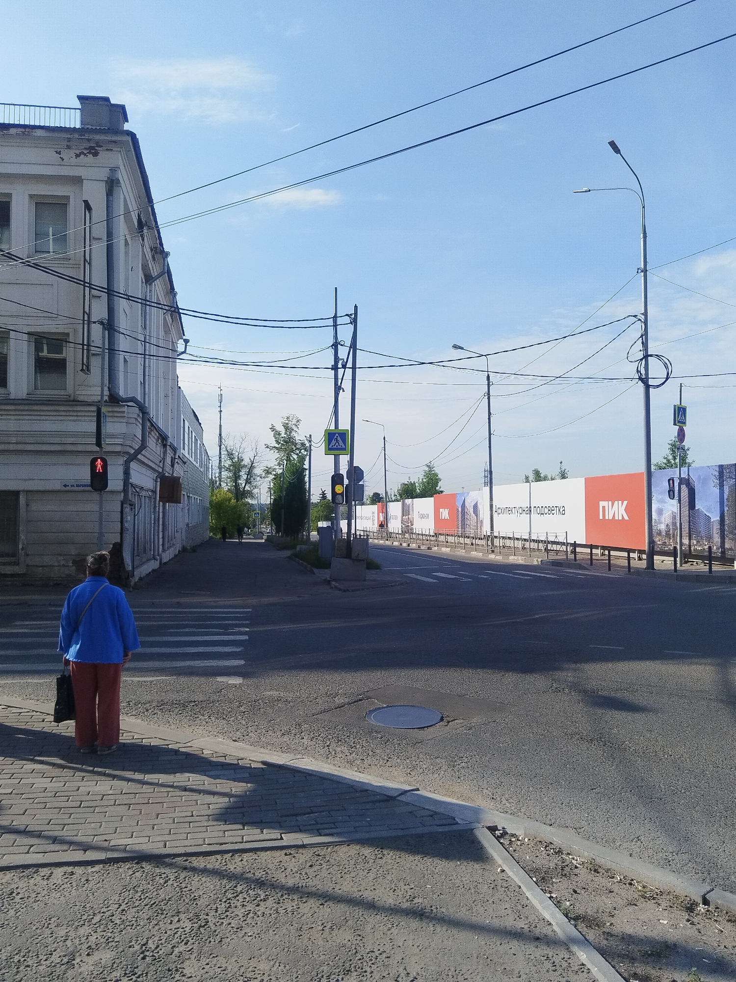 An eye-level, full shot captures a street scene on a clear day in Mytishchi, Russia. In the foreground, a person wearing a blue jacket and orange pants stands on a pedestrian crossing, facing away from the camera. To the left, a white building with a weathered facade and visible electrical wires runs along the street. A traffic light displays a red pedestrian signal. Across the street, a row of orange and white construction banners with the logo "ПИК" are visible, advertising a new development. Trees line the background, and streetlights and power lines crisscross the sky. The road surface is asphalt, with a manhole cover visible in the middle of the intersection.