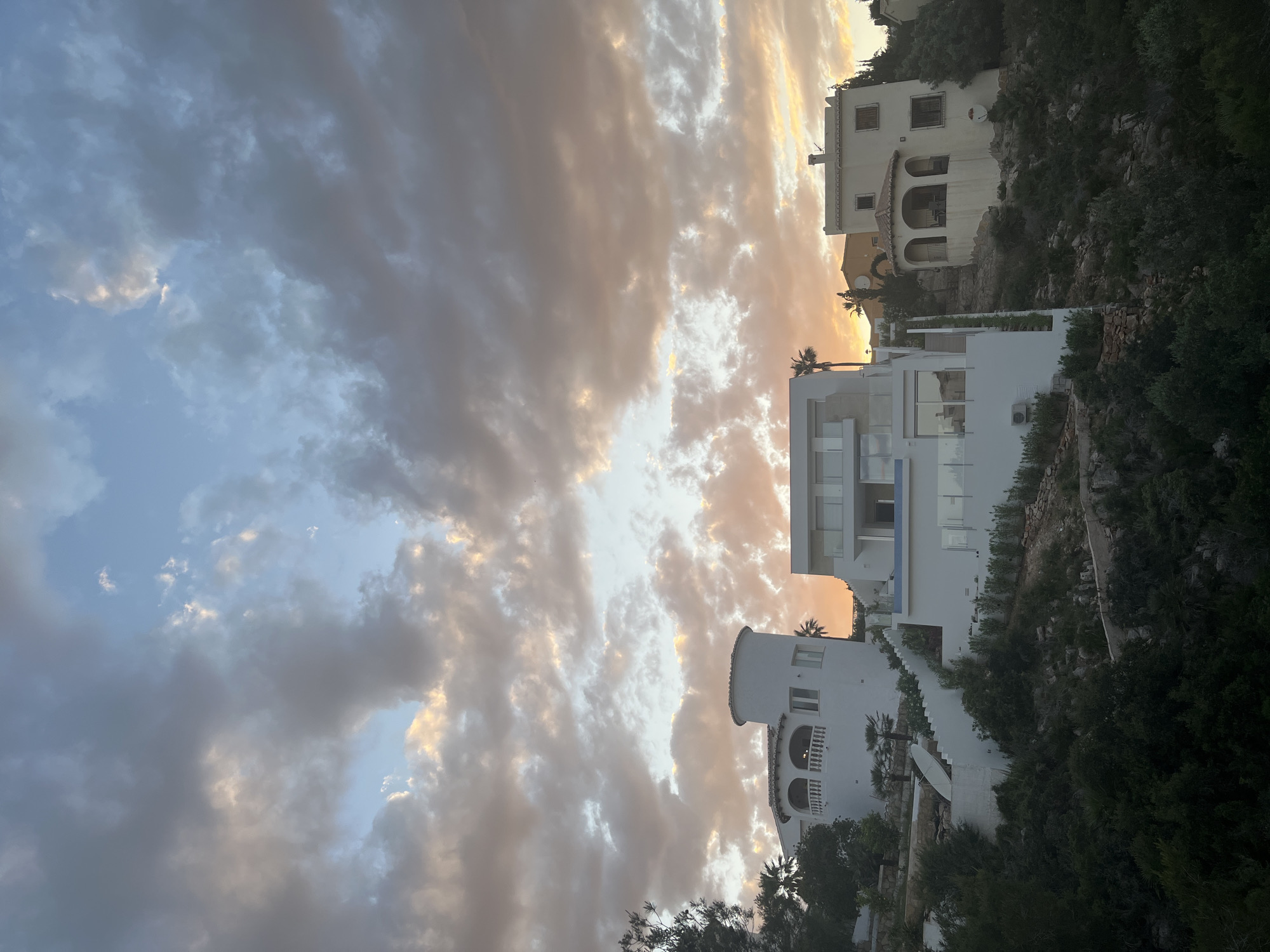 This image depicts a coastal scene in El Poble Nou de Benitatxell / Benitachell, Spain, at dusk. The sky is dominated by dramatic clouds, with patches of orange and blue light breaking through, suggesting the setting sun.
Three white buildings are visible, terraced on a hillside. The building closest to the viewer has a rounded structure with balconies. The middle building has a modern design with glass railings. The topmost building appears older, with arched openings.
The buildings are interspersed with lush green vegetation clinging to the rocky terrain. A white wall runs along the hillside, likely part of the property boundaries.