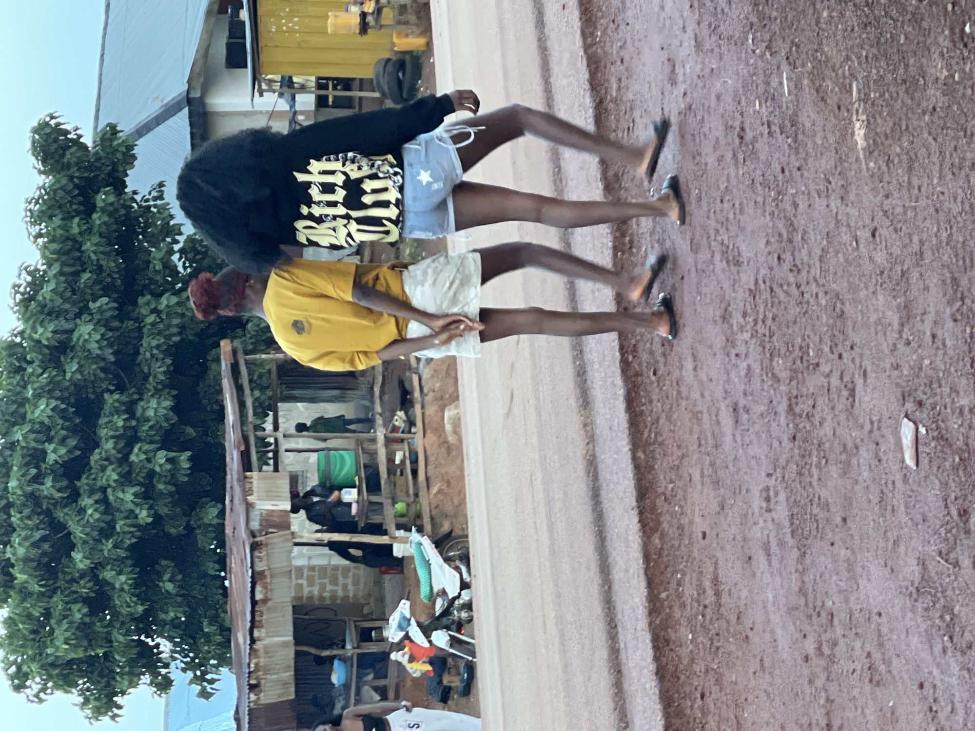 Two young women are walking away from the camera on a reddish-brown dirt road, likely in Durumi, Nigeria. The woman on the left wears a yellow t-shirt and a light-colored skirt, her hair styled in a bun. The woman on the right has on a black hoodie with "Rich Club" written on the back in yellow lettering and denim shorts adorned with a white star. Both are wearing open-toed sandals.
Behind them, a makeshift market stall with a corrugated metal roof is visible. A white motorcycle is parked near the stall. The background features lush green trees and buildings with corrugated metal roofs, characteristic of the environment in Durumi. The overall scene depicts a common street-side setting in a Nigerian city.