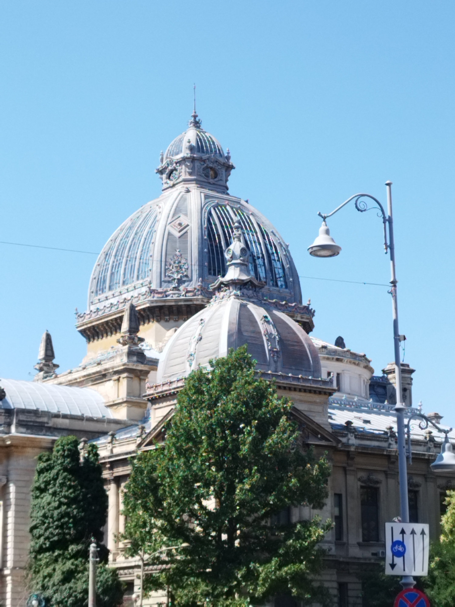 The image shows the exterior of the Cantacuzino Palace, located on Calea Victoriei in Bucharest, Romania. The palace features grand architecture, including ornate domes and intricate facades. A street lamp with a unique curved arm stands in the foreground, along with a traffic sign indicating a bicycle path and forward direction. Lush green trees partially obscure the lower sections of the building, suggesting an outdoor setting on a clear day.