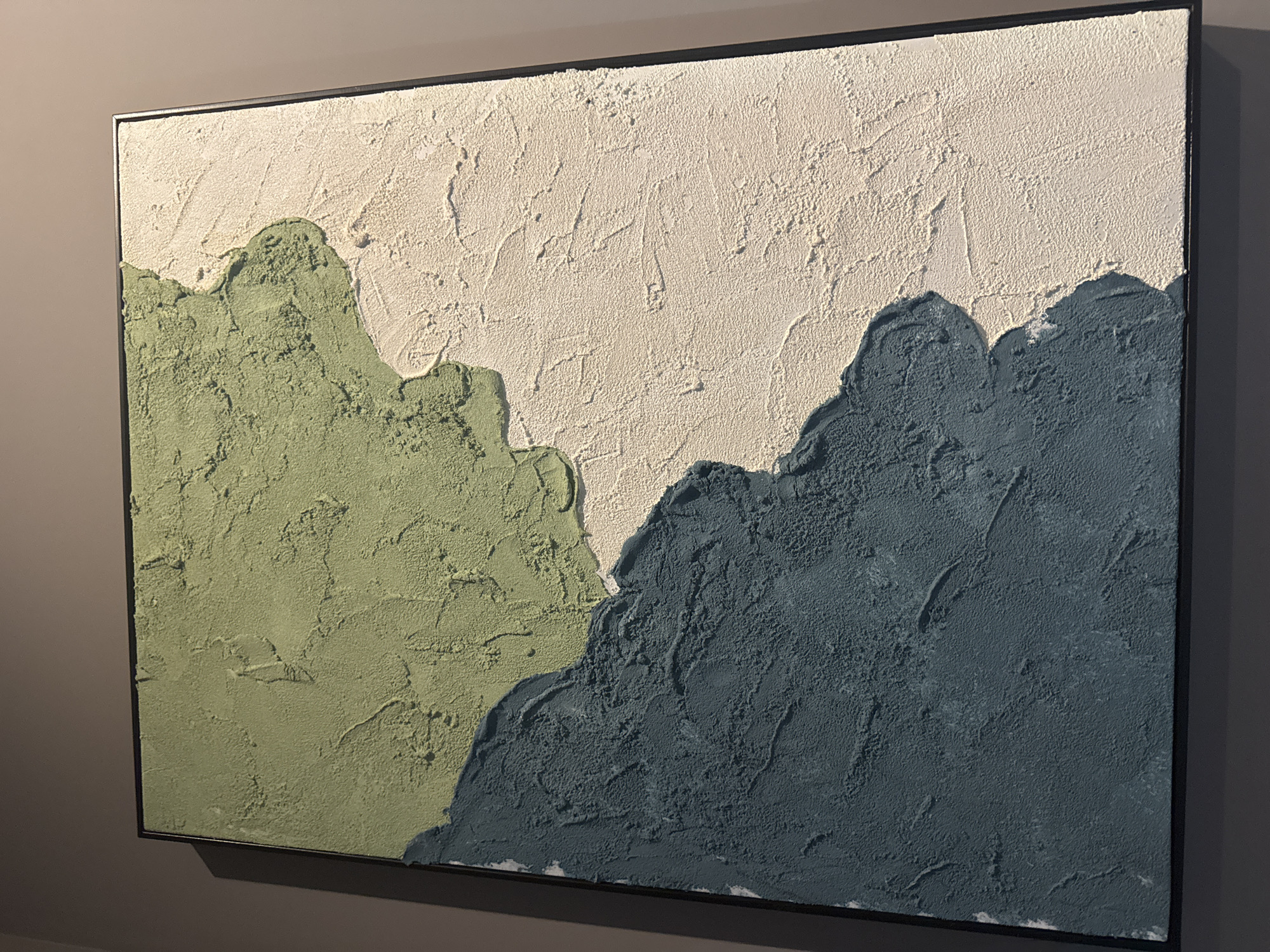 A close-up shot shows a framed, textured painting hanging on a gray wall. The painting depicts a landscape of stylized mountains. The left side of the painting features a mountain in a muted green hue, while the right side has a mountain in a deep, grayish-blue. The tops of the mountains are separated by a lighter, off-white area, possibly representing the sky or snow-capped peaks. The paint application is thick and textured, giving the artwork a tactile quality. The frame is slim and dark, contrasting with the lighter tones of the painting and the surrounding wall.