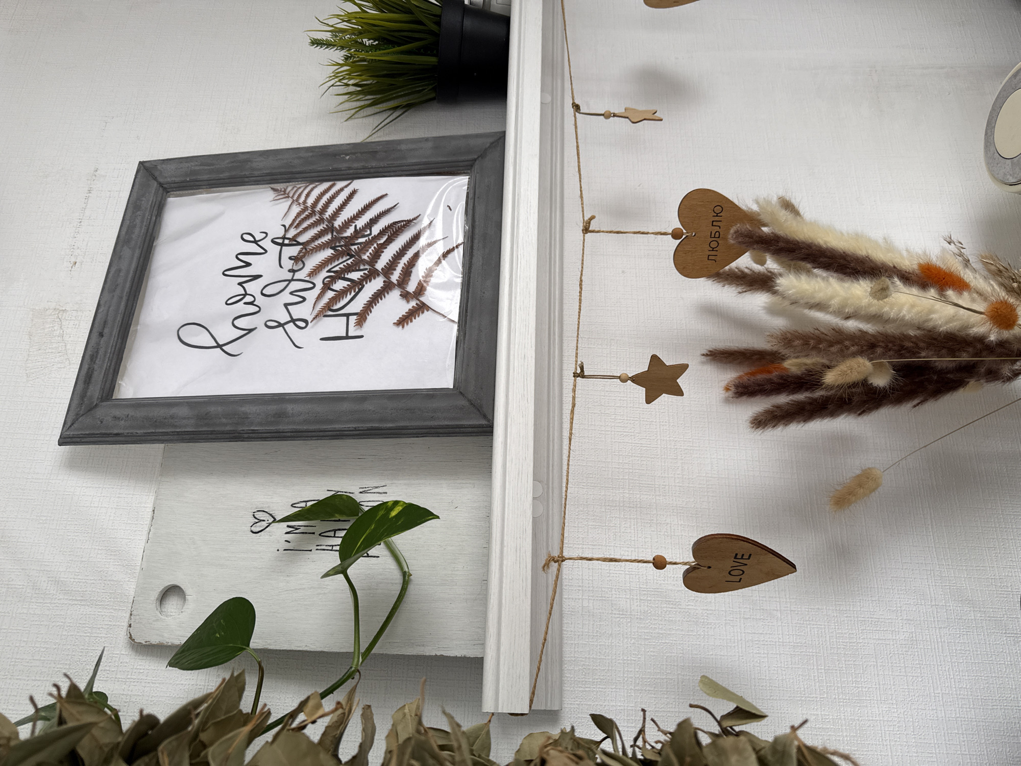 The image shows a wall decorated with various objects. At the top left, there's a framed picture featuring a dried fern and lettering. Above it, part of a potted green plant is visible. Below the framed picture, there's a small white wooden board with a leaf design, and a green vine with leaves is trailing across it. To the right, there's a light-colored wooden panel with hanging decorations including wooden hearts and stars. Dried floral arrangements extend from behind the panel. The bottom of the image is obscured by dried leaves and stems. The wall is textured and white. The image was taken in Chaikovsky, Russia, as evidenced by the Cyrillic text "ЛЮБЛЮ" on one of the wooden heart decorations.