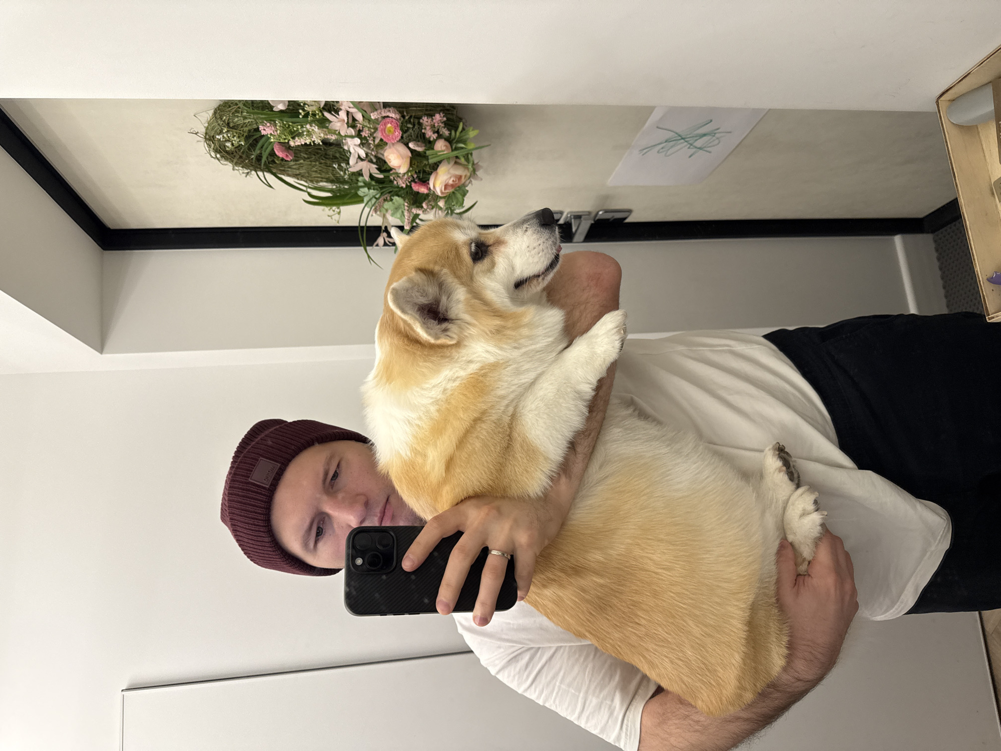 A man wearing a maroon beanie, white t-shirt, and black pants is holding a corgi dog in his arms. He is taking a selfie with a black iPhone. The man is wearing a silver wedding ring on his left ring finger. Behind him, there is a flower arrangement on a mirrored wall, and a white piece of paper with a green scribble is taped to the mirror. A wooden box is also visible near the top right corner of the image. The location is unknown.