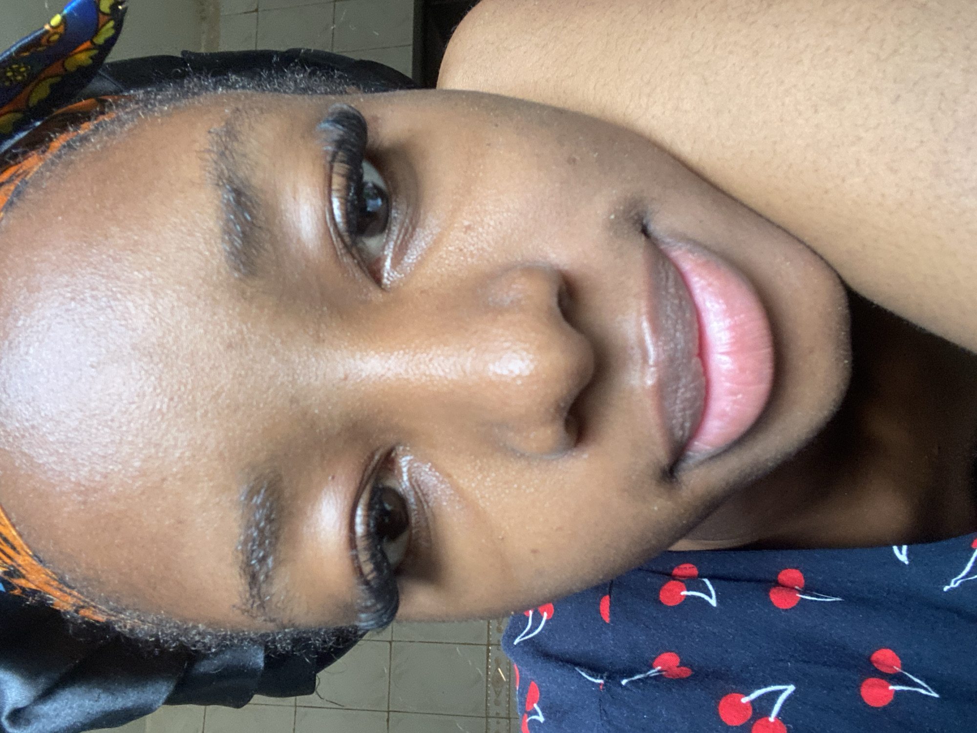 A close-up portrait of a young individual with dark skin, likely taken in Jos, Nigeria. The individual's head is partially covered by a black satin-like cap, with a segment of an orange, blue, and black patterned fabric visible along the hairline. Both eyes, featuring prominent dark eyelashes, are open and directed towards the camera. The eyebrows are dark and naturally full. The nose is broad, and the full, light pink lips are slightly parted, forming a gentle smile. The skin exhibits natural texture and a subtle sheen, particularly on the forehead and cheeks. The individual wears a dark blue top with a repeating pattern of red cherries and white stems, visible on the left side of the frame. The lower right portion of the image shows skin texture, suggesting the individual's face is resting against an arm or shoulder. In the upper left background, a light-colored tiled wall with visible grout lines is present.