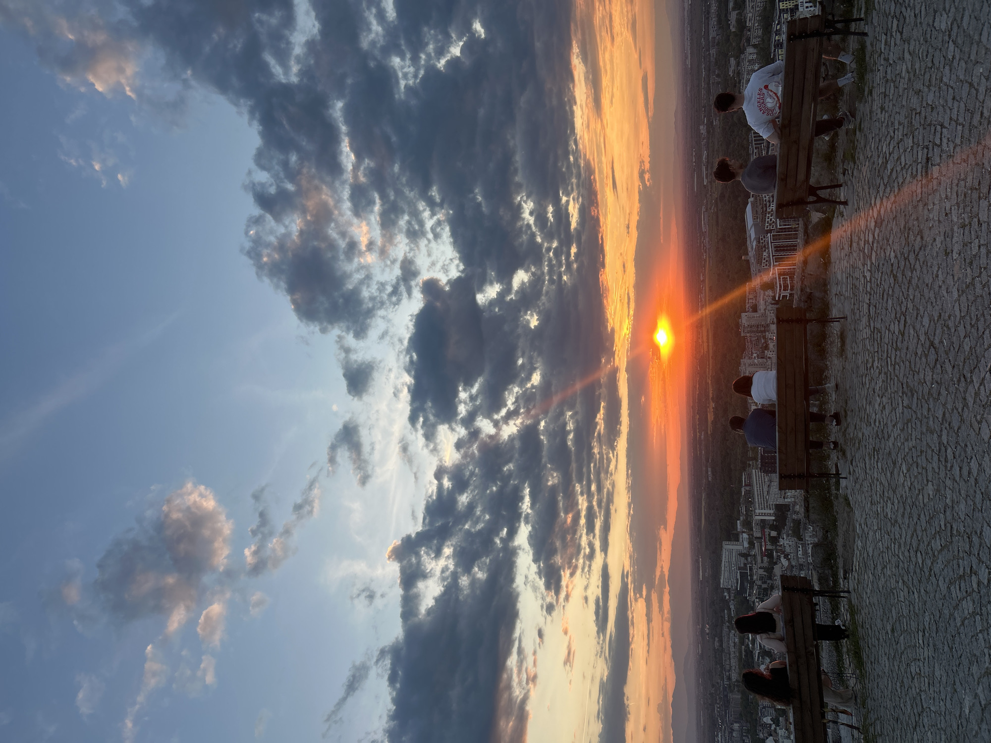 The image depicts a sunset over the city of Plovdiv, Bulgaria. The sun is setting with an orange glow on the right side of the image. Above, there is a dramatic sky with a mix of blue sky and dark, heavy clouds. Several people are sitting on benches on a stone-paved surface, facing the sunset. The benches are made of wood. A city skyline with buildings is visible beneath the sunset's glow. The image is oriented sideways.