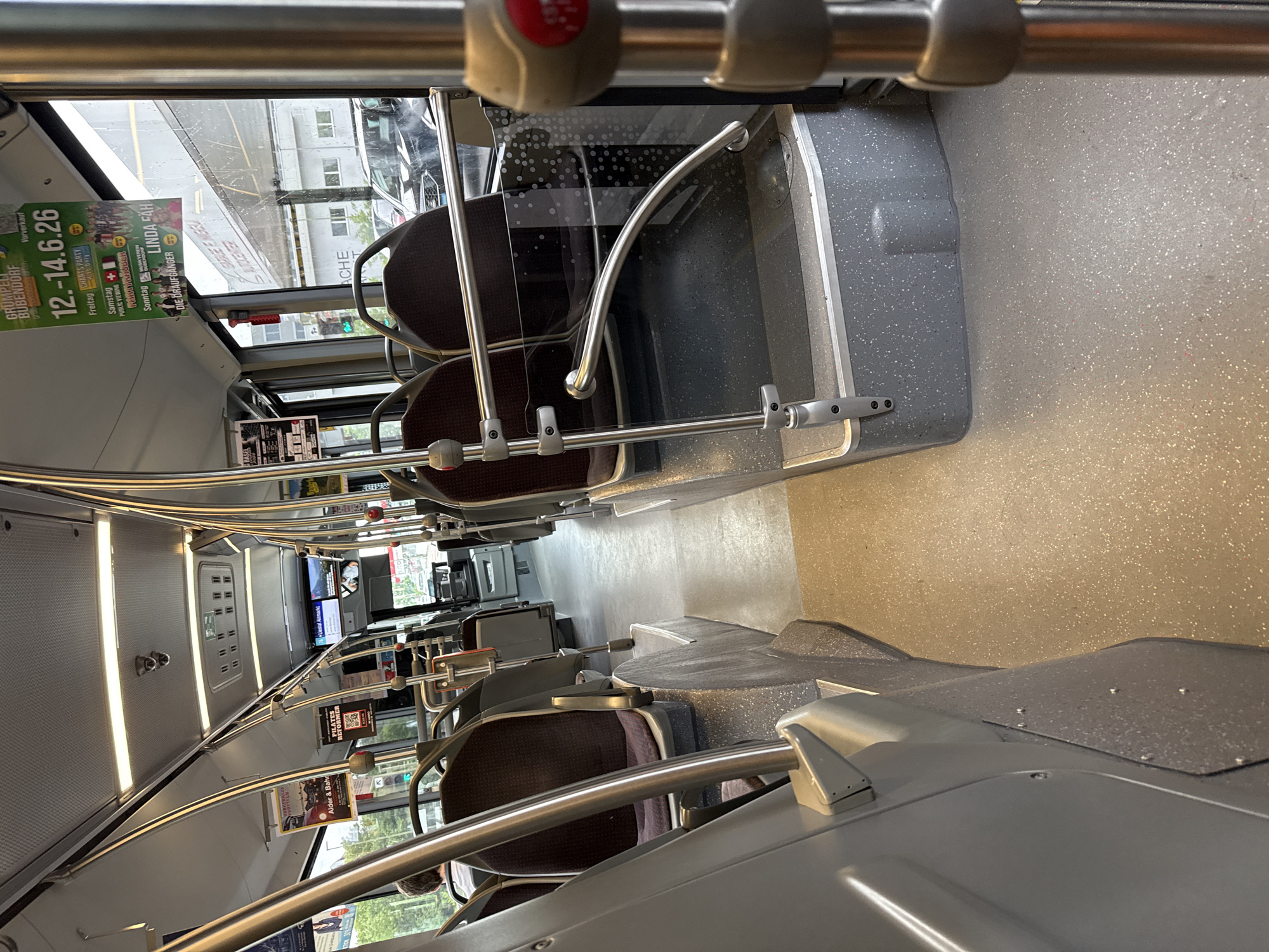 The image shows the interior of a public bus in Liestal, Switzerland. The bus is mostly empty, with rows of brown padded seats along the sides of the aisle. Silver metal handrails run along the seats and overhead. Advertisements are visible on the windows and seat backs. Outside the windows, buildings and a street scene can be seen.