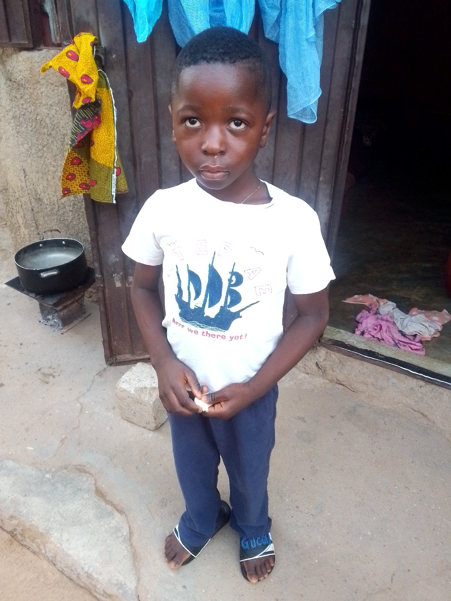A young Black boy stands in front of a wooden doorway. He is wearing a white t-shirt with a blue graphic of a sailboat and the text "Arrr we there yet!", dark blue pants, and black and white slides. He is holding a small white object in his hands. To the left of the boy, a black pot with boiling water sits on a makeshift stove. In the background, a colorful patterned cloth hangs on the wall, and a pile of pink and gray fabric is on the ground near the doorway. The ground is concrete with some cracks and a few scattered stones. The overall scene suggests an outdoor living space, possibly in a less developed urban area.