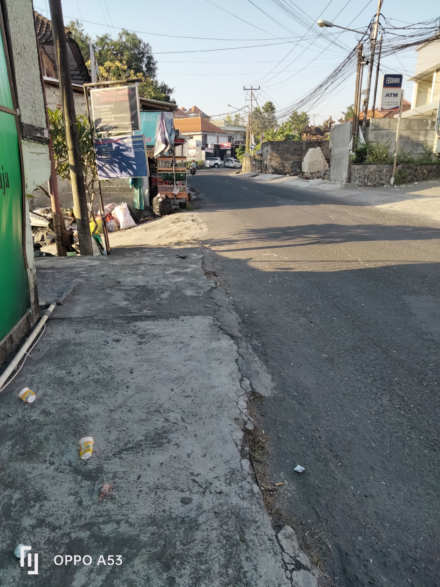 This is a daytime, eye-level, outdoor photograph of a street scene in Denpasar, Indonesia. The image is dominated by a paved road, which curves to the right, leading into the distance. On the left side of the road, there is a concrete sidewalk with several discarded paper cups and a small piece of debris. Adjacent to the sidewalk is a building with a rough, light-colored brick wall. Several signs are attached to a pole in front of this building, advertising "BENGKEL MOBIL ARTHA JAYA" (Artha Jaya Car Workshop) and other services. Further down the street on the right, a BRI ATM machine is visible on a pole. Buildings line both sides of the road, with visible power lines strung overhead. The environment appears to be an urban or semi-urban area with a clear sky. There are no people actively engaged in any visible interactions in the immediate foreground.