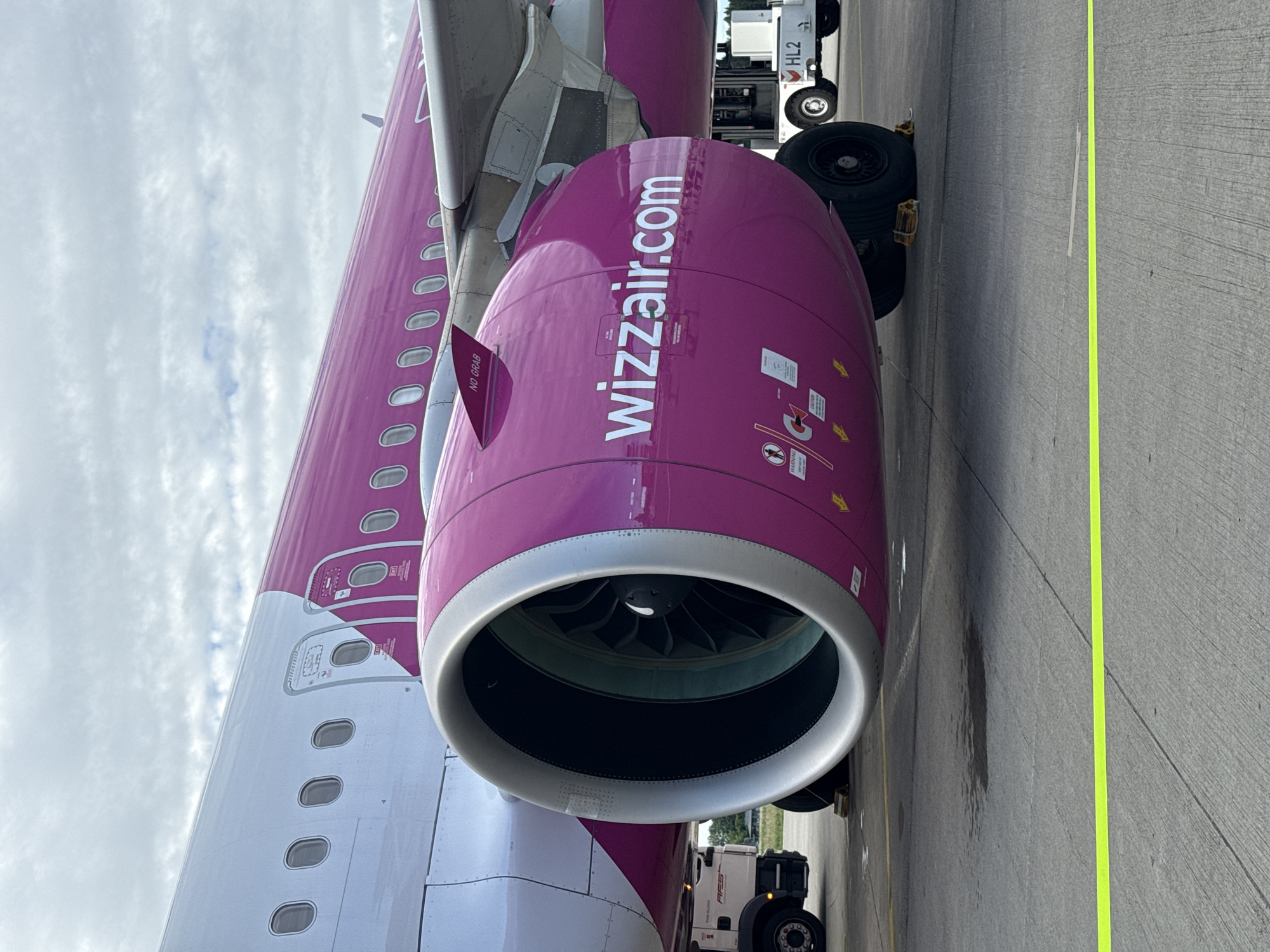 An airplane engine, painted magenta with "wizzair.com" written on it in white letters, is visible in the foreground. The engine is large and cylindrical with visible fan blades inside. The fuselage of the airplane, white with a magenta stripe, extends to the left. Several windows are visible on the fuselage. A yellow caution tape runs across the concrete tarmac in front of the plane. In the background, a white utility vehicle with "HL2" on its side is parked to the right of the engine. To the left of the engine, a service truck is parked. The sky is cloudy. The scene appears to be at an airport in Künersberg, Germany.