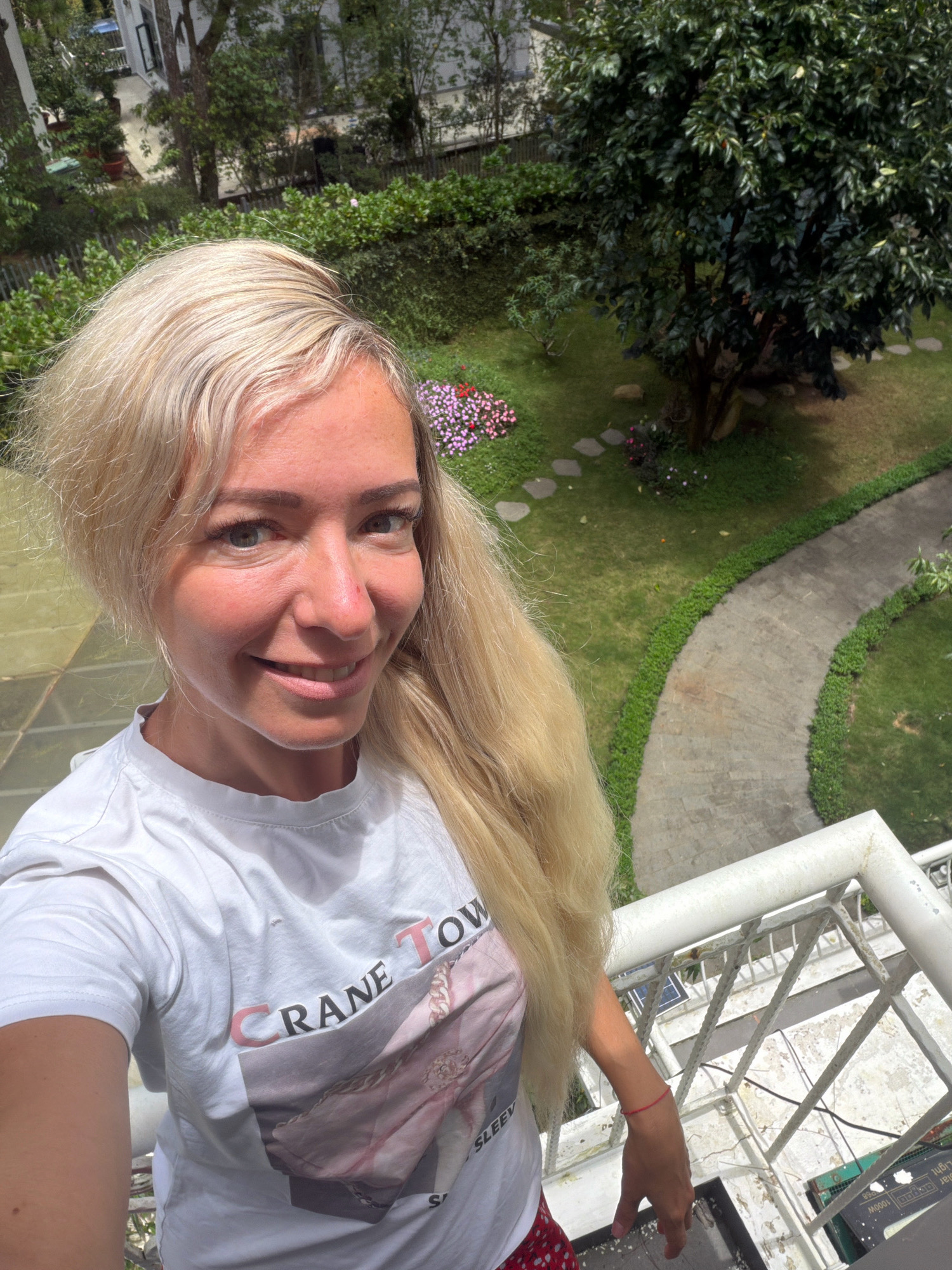 A woman with blonde hair is taking a selfie on a balcony overlooking a garden. She is wearing a white t-shirt with a graphic print and red and white polka dot shorts. The garden features a winding pathway, green grass, trees, and flower beds. The woman is smiling and looking at the camera. The balcony has a white railing and some discarded items below, including what appears to be solar panels. The background shows buildings and lush greenery, indicative of a tropical or subtropical climate. The location is identified as Xuân Trường - Đà Lạt, Vietnam, a city known for its cool climate, pine forests, and French colonial architecture.