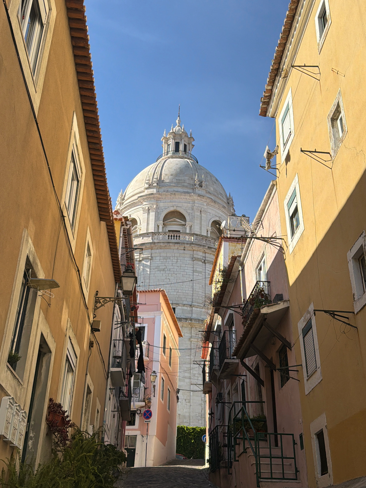This is a low-angle, medium shot of a narrow cobblestone street in Lisbon, Portugal, leading towards a large, domed white building, likely the Pantheon of Lisbon. The street is flanked by two- and three-story buildings with ochre and pastel pink facades. The building on the left is painted ochre and features traditional Portuguese tilework on its roofline, while the building on the right is a pale yellow with a prominent shadow cast across it by the sun. Both buildings have several windows with shutters, and some feature wrought-iron balconies. Laundry hangs from lines strung between some of the buildings. A street lamp is mounted on the ochre building, and a traffic sign is visible in the distance. The sky above is clear and blue. No people are visible in the image.