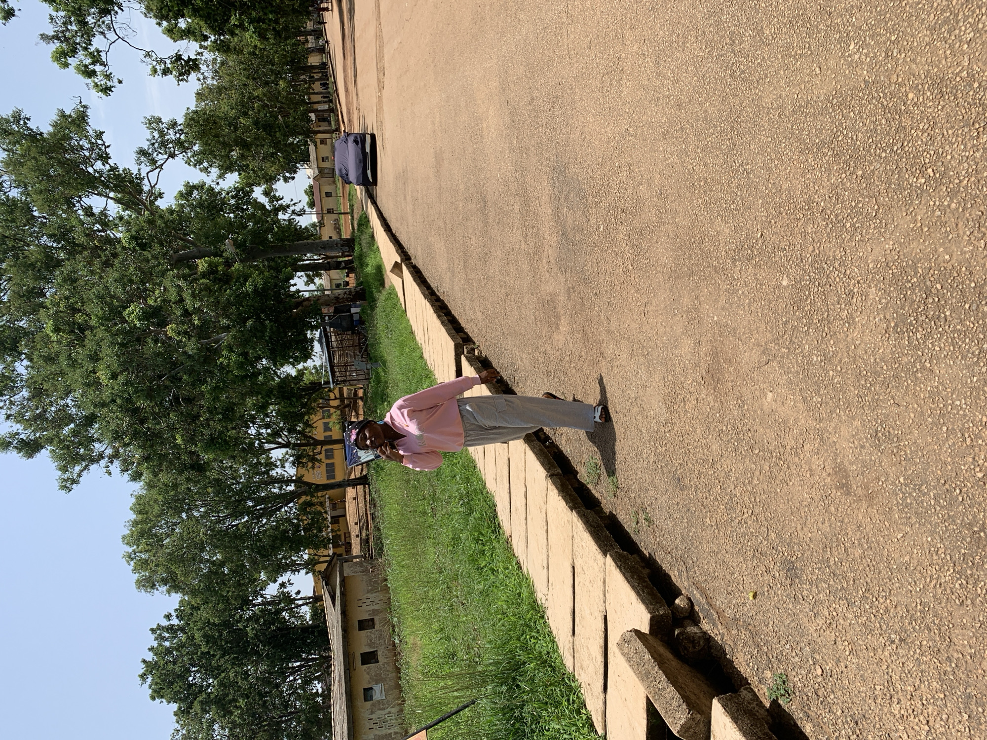 A woman in a pink long-sleeved shirt and grey pants walks along a paved walkway next to a dirt road. She is holding a phone to her ear. The walkway is made of large concrete slabs, some of which appear to be damaged and dislodged. Lush green grass grows on the other side of the walkway, and a building with a yellow facade can be seen in the background. Trees line the side of the road, and a car covered in a dark tarp is parked further down. The sky is clear and blue. This scene is located on the grounds of the Federal University of Agriculture, Abeokuta (FUNAAB), in Abeokuta, Ogun State, Nigeria.