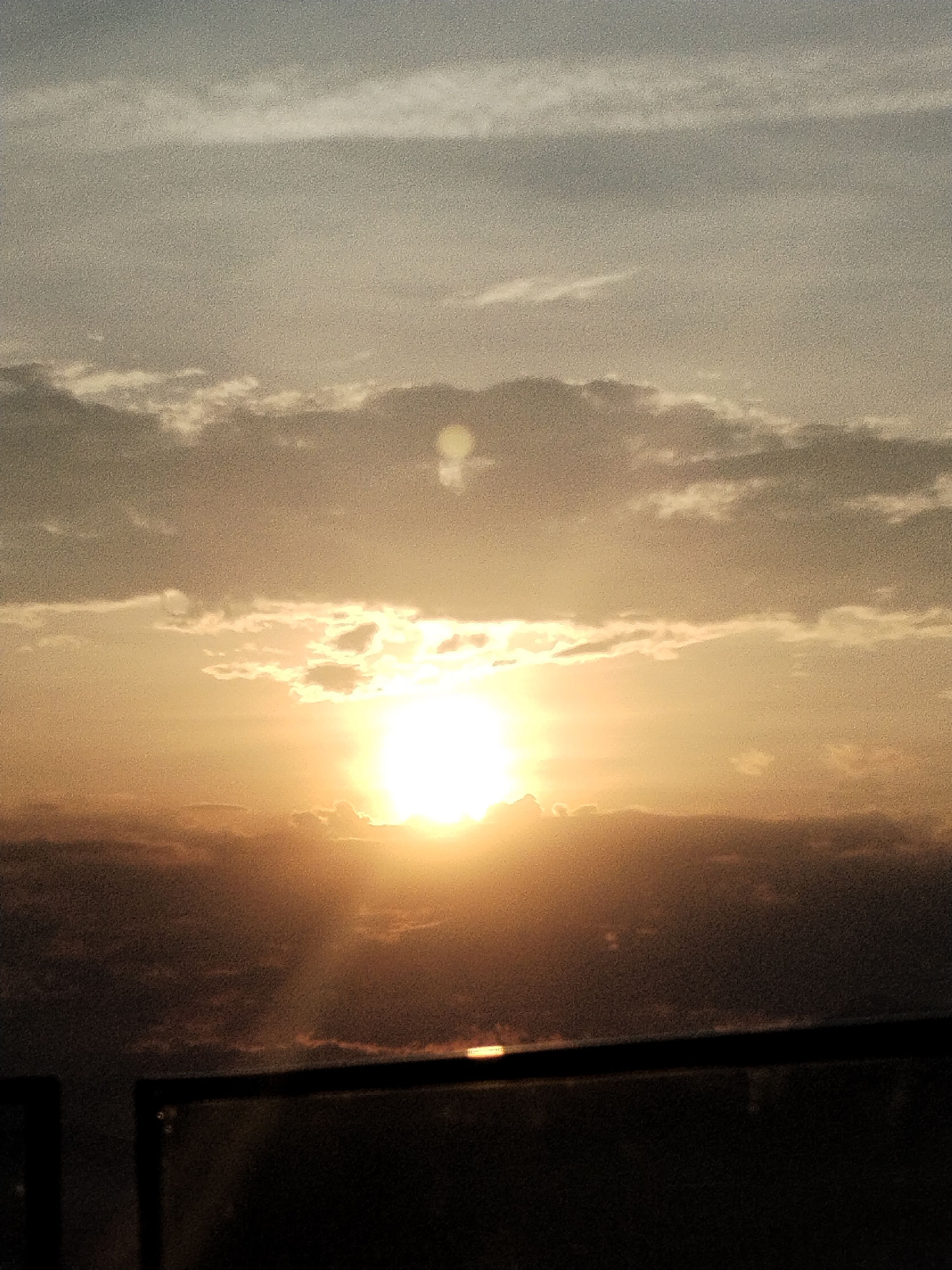 A hazy, golden sunset is captured behind a silhouette of clouds and a dark foreground structure, possibly a railing. The sun is a bright, almost blinding orb in the center of the frame, with rays of light emanating outwards. The sky is a gradient of pale blue and soft orange, with scattered clouds catching the warm light. The foreground appears to be a dark, undefined structure, possibly a balcony or rooftop, obscuring the ground. The image suggests an elevated viewpoint overlooking a landscape or city, but the details are indistinct due to the low light and atmospheric conditions. The location is specified as Nha Trang Ward, Vietnam.