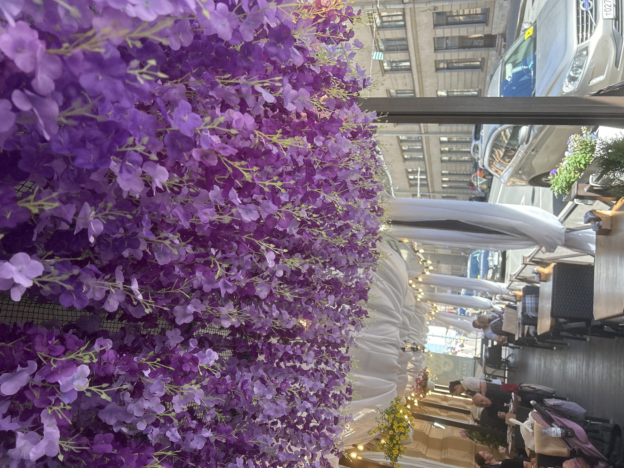 An outdoor seating area of a restaurant is decorated with abundant purple flower garlands hanging from the ceiling. White drapes and string lights are also visible, creating a festive atmosphere. Several tables and chairs are arranged for patrons. In the background, a street scene with buildings and cars can be observed. A few people are seated at tables, engaged in dining or conversation. The overall impression is a well-decorated, lively outdoor dining space in an urban setting.