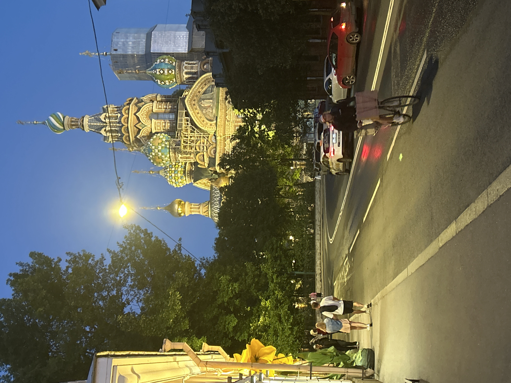 An outdoor, eye-level, vertical shot captures a street scene at dusk in Unknown. The sky is a deep, twilight blue. In the background, the intricately decorated Church of the Savior on Spilled Blood, a landmark in Saint Petersburg, Russia, is illuminated. The church features multiple domes of various colors, including green, blue, and gold, and ornate architectural details. Adjacent to the church, a modern glass-and-steel building stands, creating a contrast between old and new architecture.
In the foreground, a tree-lined sidewalk runs along the left side of the frame. A couple is walking away from the camera on the sidewalk; the woman wears a denim jacket and a light-colored skirt, and the man wears a white t-shirt and black shorts. To their left, a building with yellow siding is visible, adorned with a large, artificial yellow flower. A green trash receptacle is placed near the sidewalk.
On the street, a cyclist, wearing a dark jacket and a baseball cap, rides a bicycle equipped with a large basket in the foreground, moving towards the right of the frame. Several cars are on the road, with headlights and taillights indicating traffic. The street is paved and marked with white lines. The overall atmosphere suggests a bustling urban environment transitioning into evening.