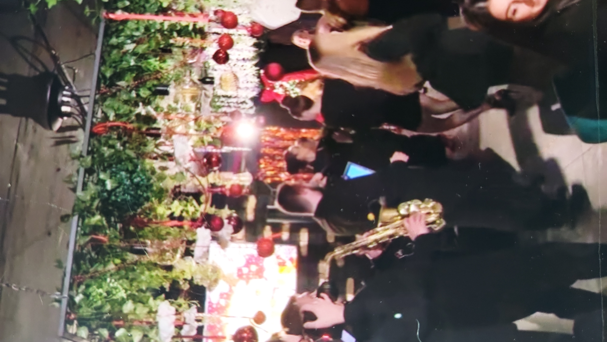 In an indoor setting, possibly a venue, a live band performs. A saxophonist, positioned on the left and facing left, plays a golden saxophone. They are dressed in black attire. To the right of the saxophonist, other band members in black attire are visible, including a person with their back to the camera. In the background, a group of people are dancing and socializing. The environment is decorated with artificial greenery hanging from above, interspersed with red ornaments and red ribbons, suggesting a festive or holiday atmosphere. A large screen displaying colorful patterns is visible behind the performers.