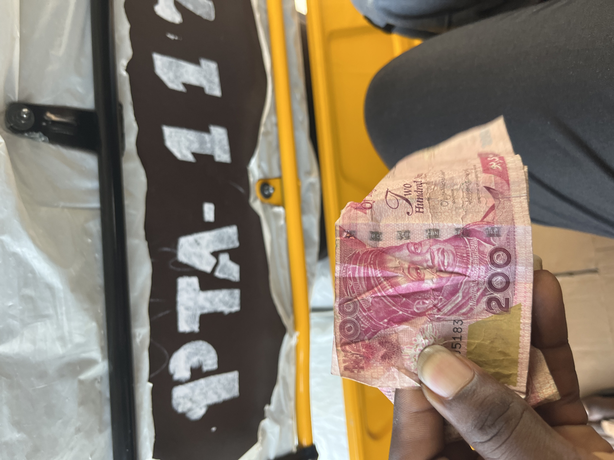 This media file shows a close-up of a hand holding Nigerian currency, specifically a 200 Naira note. The background suggests an outdoor or vehicle-based setting, with a glimpse of yellow and dark surfaces, possibly part of a trishaw or similar transport. A sign with white lettering on a dark background, partially visible, reads "PTA-117". The currency is pink and features a portrait of a man, likely Alhaji Sir Ahmadu Bello, along with the text "Two Hundred" and the numeral "200". The hand holding the money appears to be that of a person of color. The scene is lit, possibly by daylight. The overall impression is a transaction or payment taking place in a casual, everyday environment within Nigeria, as suggested by the currency and location context.