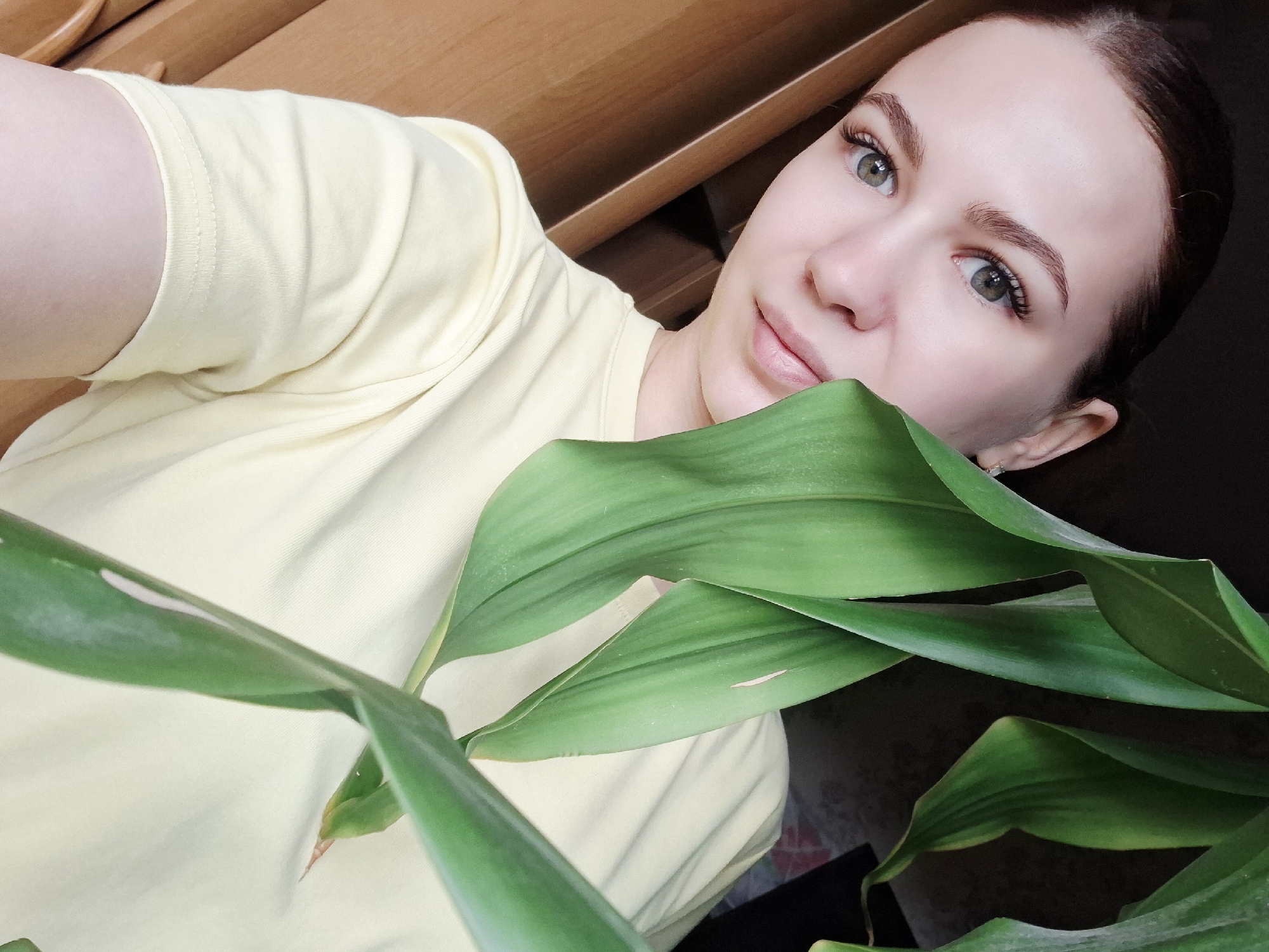 A woman with fair skin, green eyes, and brown hair pulled back is positioned in front of a green leafy plant. She wears a yellow t-shirt. The background includes what appears to be a wooden cabinet or shelf. The location is described as Nizhnyaya Gar', Russia.