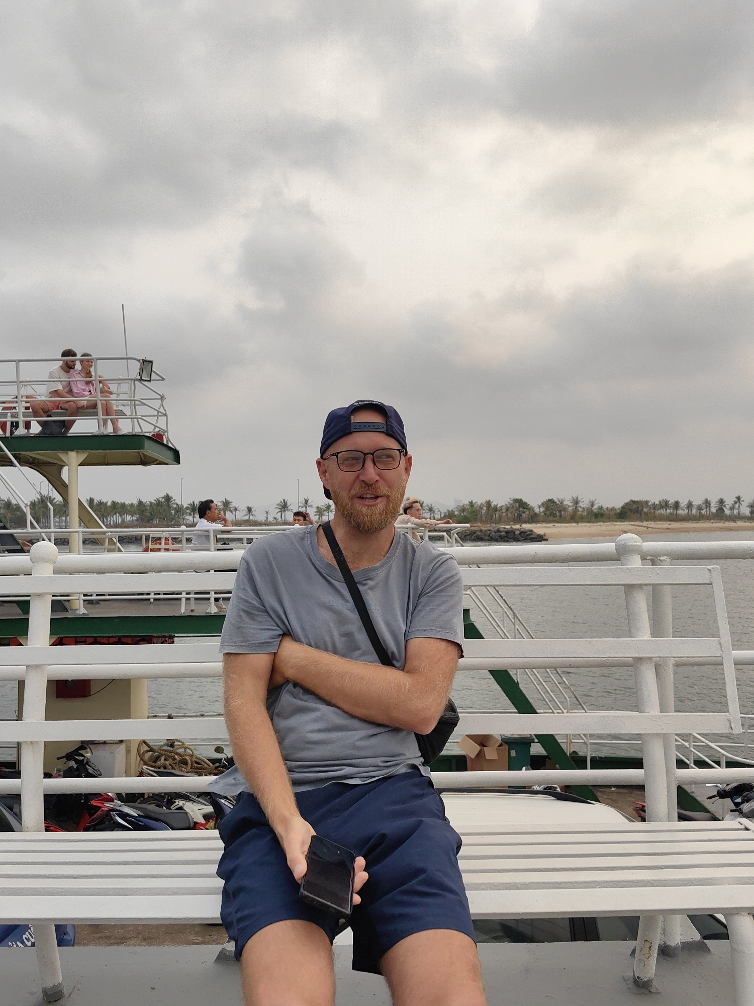 The image shows a man seated on a bench on a ferry. He is wearing a grey t-shirt, blue shorts, and a blue baseball cap. He has a beard and is wearing glasses. He is holding a black smartphone in his left hand, and his arms are crossed. Behind him, there is a white railing and a view of the sea, with a coastline visible in the distance featuring palm trees. Other passengers and portions of the ferry's upper levels are also visible. The location appears to be in Tuan Chau Ward, Vietnam.