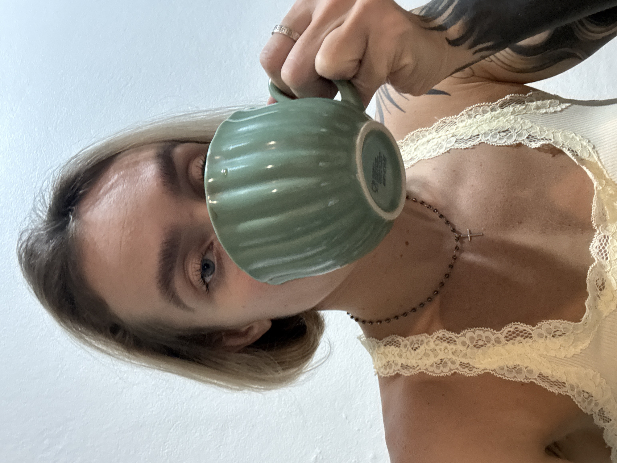 A fair-skinned person with blonde hair is holding a green, ridged mug to their lips as if drinking from it. The person is wearing a cream-colored top with lace trim along the neckline and a delicate necklace with a cross pendant. They also have a ring on one finger and a tattoo on their upper arm. The background is a plain white wall. There is no verifiable information available to indicate this photograph was taken in Karon, Thailand.