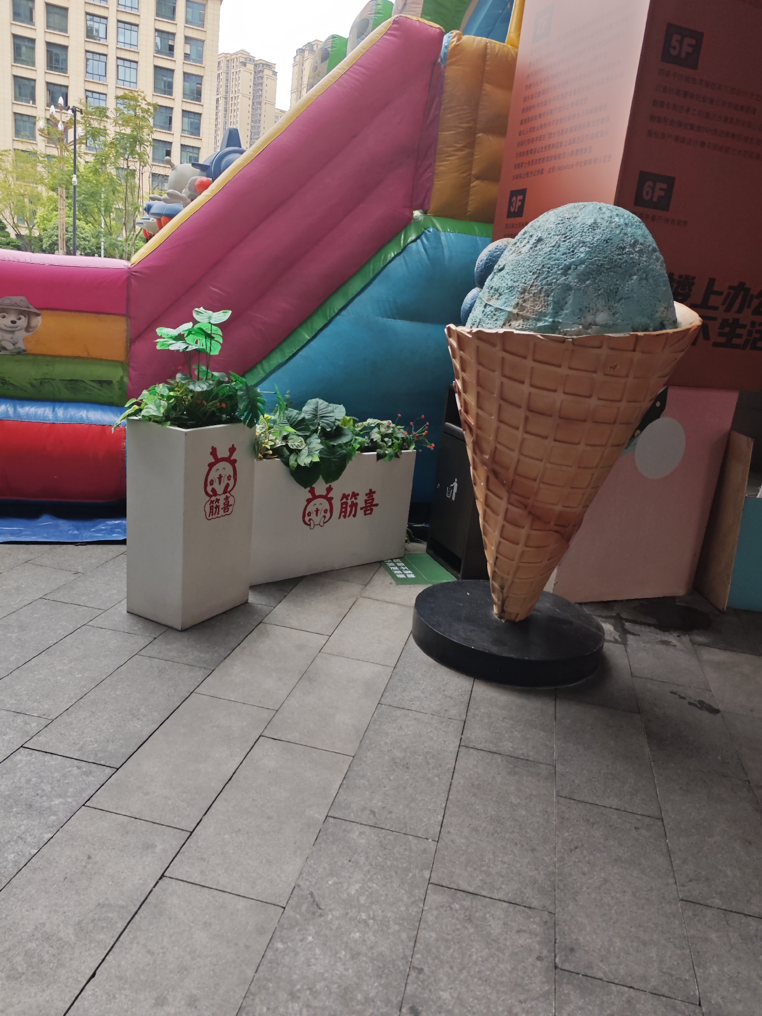 The image shows a scene in what appears to be an outdoor commercial area in Liangjiang, China. The foreground contains two white rectangular planters filled with greenery. Next to the planters stands an oversized sculpture of an ice cream cone, featuring a blue ice cream scoop in a brown waffle cone. Behind these objects is a multi-colored inflatable structure, possibly a bouncy castle. In the background, there's a tall building with multiple floors, suggesting an urban setting. To the right of the ice cream sculpture are large panels with Chinese writing, possibly advertisements or building directories. The ground is paved with grey tiles.