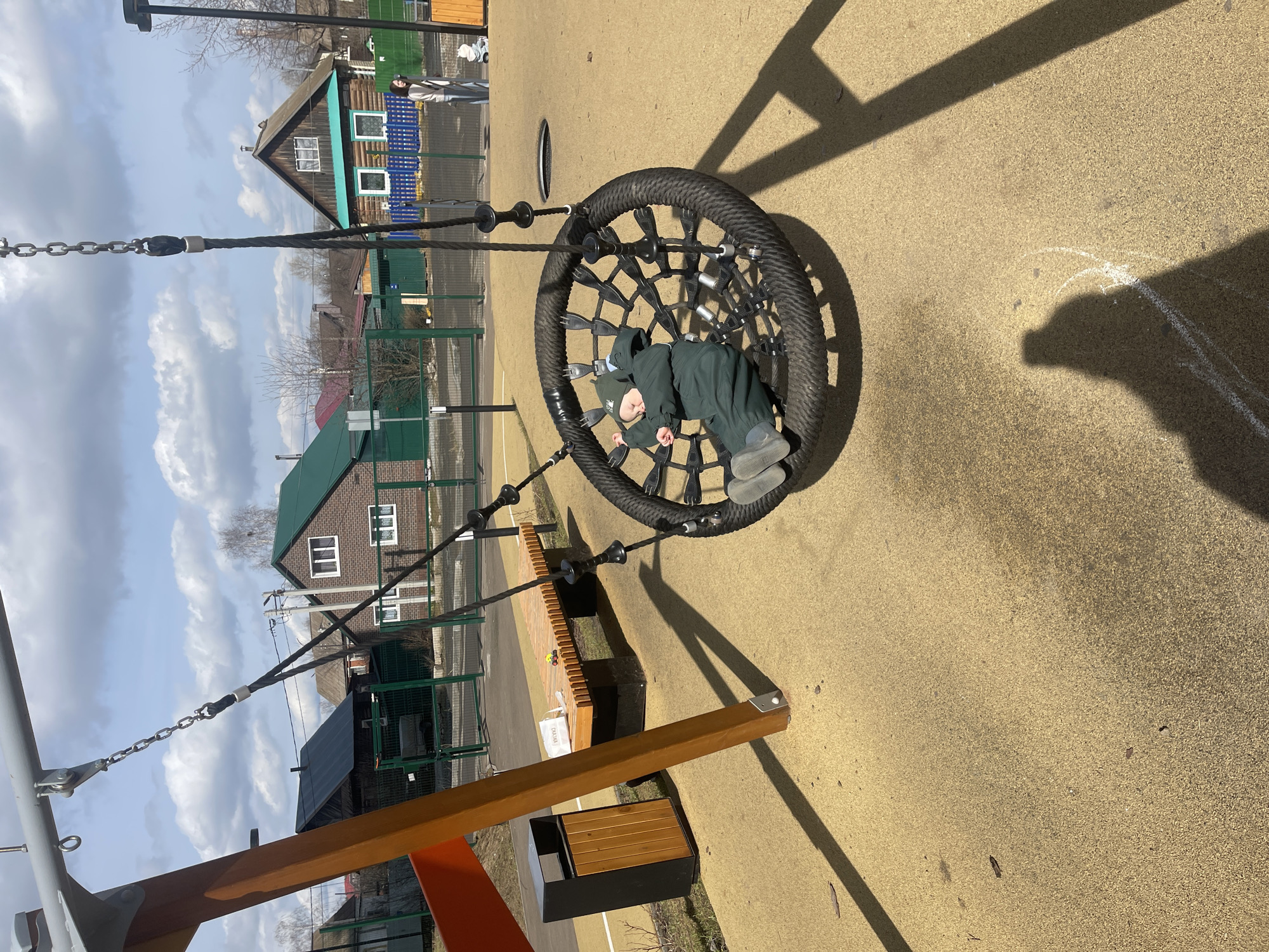 A young child dressed in a dark green snowsuit and matching hat lies in a circular net swing at a playground. The swing is suspended by thick black ropes connected to a wooden frame. The playground has a soft, granular, yellow-beige surface. A wooden bench and a trash bin are visible nearby. In the background, houses with green and brown painted exteriors are partially visible under a partly cloudy sky. The setting is Izhevsk, Russia. A person stands in the background.