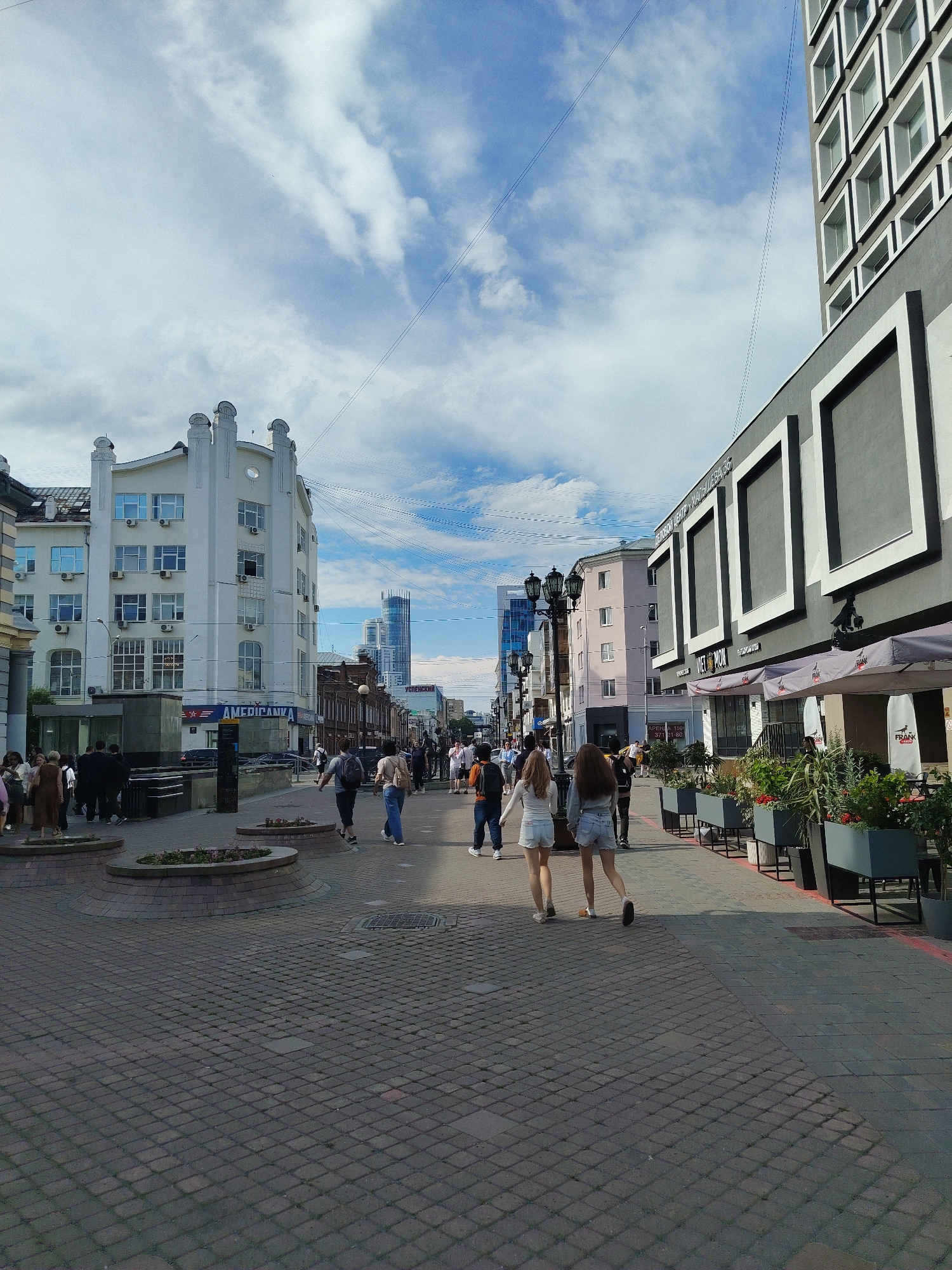 The image captures a street scene in Yekaterinburg, Russia, likely on a sunny day with scattered clouds. The perspective is from ground level, looking down a pedestrian walkway. To the left, a prominent white building with ornate architectural details and numerous windows stands out. Below this building, a street-level entrance with signage is visible, including the word "AMERICANKA." Further down the street, more buildings of varying architectural styles and heights are present, including a modern skyscraper in the distance.
On the right side of the walkway, a contemporary building with large, dark rectangular panels is visible, featuring awnings from a business, possibly a cafe or restaurant. Planters with greenery line this side of the walkway, and outdoor seating is implied.
The walkway itself is paved with dark cobblestones arranged in a diamond pattern. Several individuals are walking along the path. In the foreground, two young women in casual summer clothing, shorts and tops, are walking away from the camera. Other pedestrians, a mix of adults and some in groups, are scattered throughout the scene, engaged in walking or standing. Streetlights are visible along the walkway. The overall impression is of a busy urban commercial or pedestrian area.