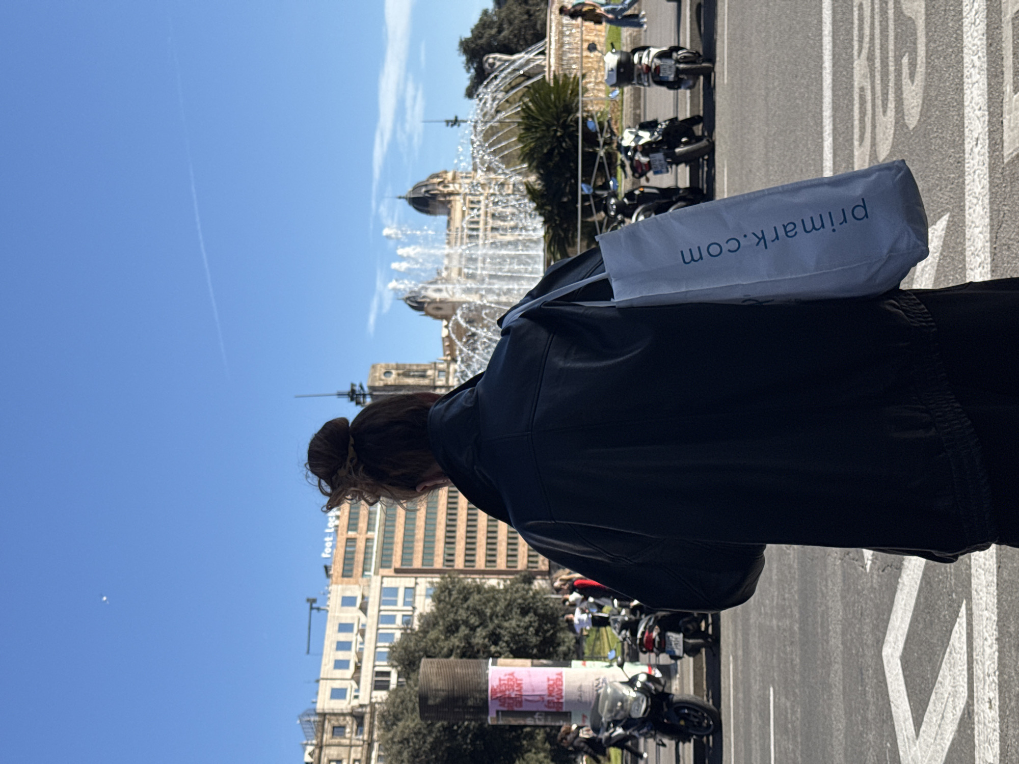 A woman with brown hair tied in a bun stands on a street in Barcelona, Spain. She wears a black jacket and carries a white bag with "primark.com" printed on it. Behind her is the Magic Fountain of Montjuïc, a large fountain complex, with its intricate architecture and water display visible. In the background, there are buildings and clear blue sky. The street shows the word "BUS" and motorcycles parked.