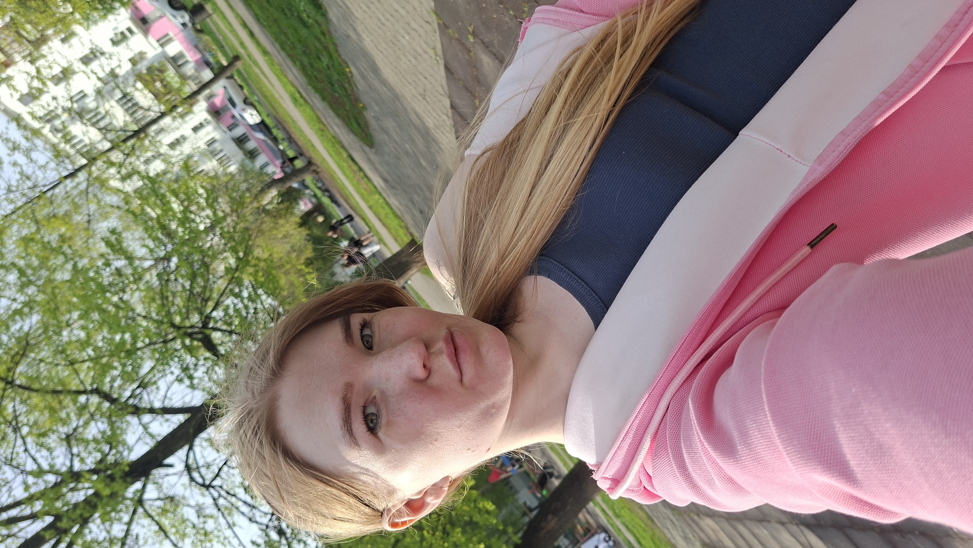 A woman with blonde hair and fair skin is positioned in the foreground. She is wearing a dark blue tank top and a pink and white hoodie. The background includes trees with green leaves, suggesting a park or outdoor area. In the distance, a light-colored building with pink accents is visible, along with a paved area. The location appears to be in an urban environment, potentially in Chaikovsky, Russia. A group of people are gathered near the tree in the background.